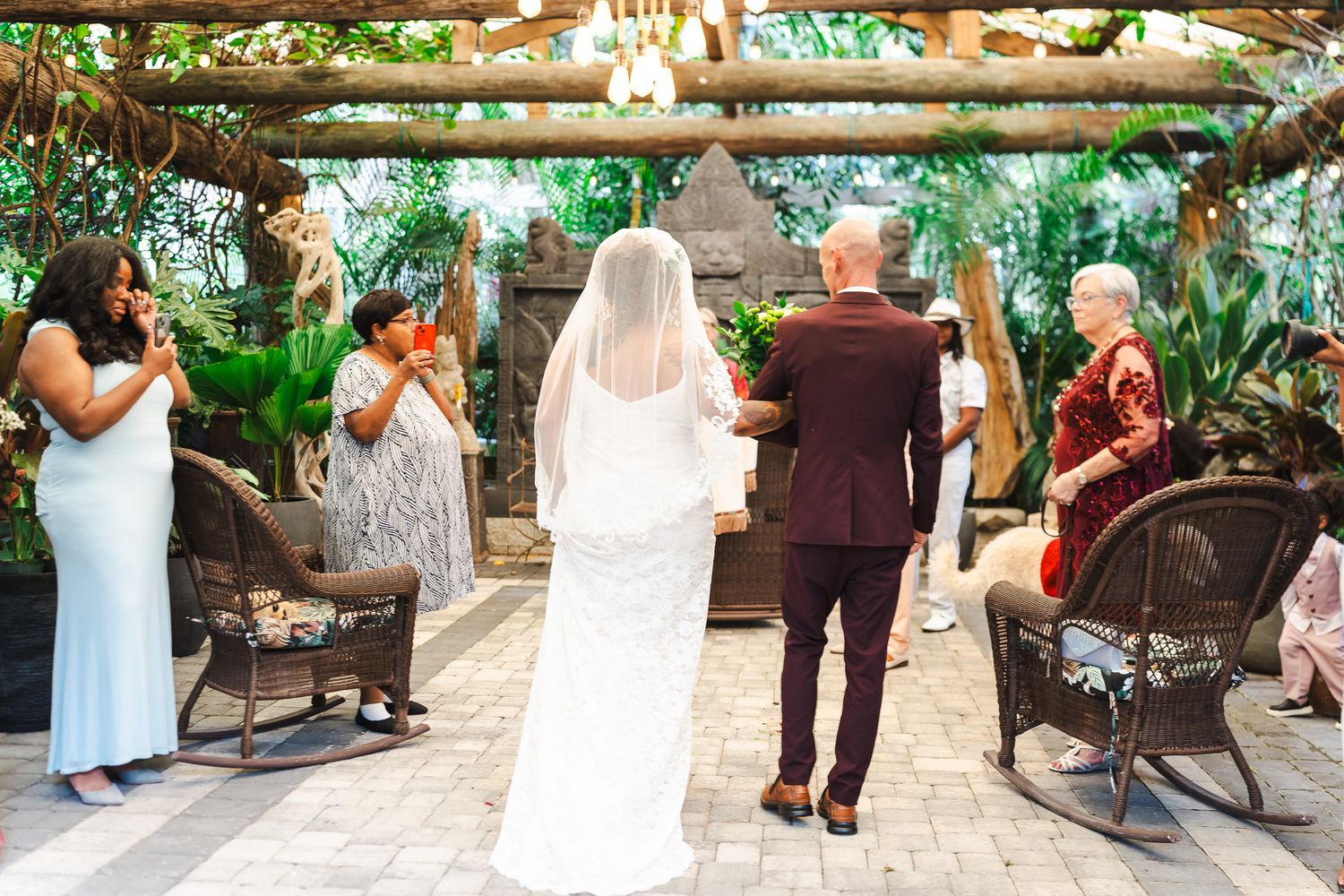 Wedding ceremony in tropical garden with bride, groom, and officiant under a wooden pergola with guests.