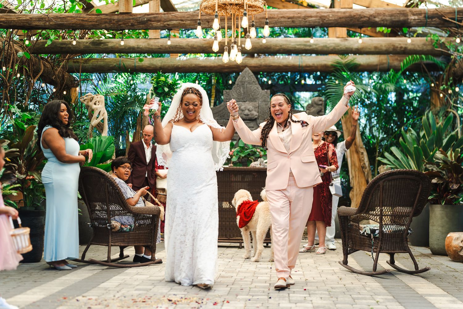 Two brides celebrate joyfully as they exit their garden wedding ceremony surrounded by guests.