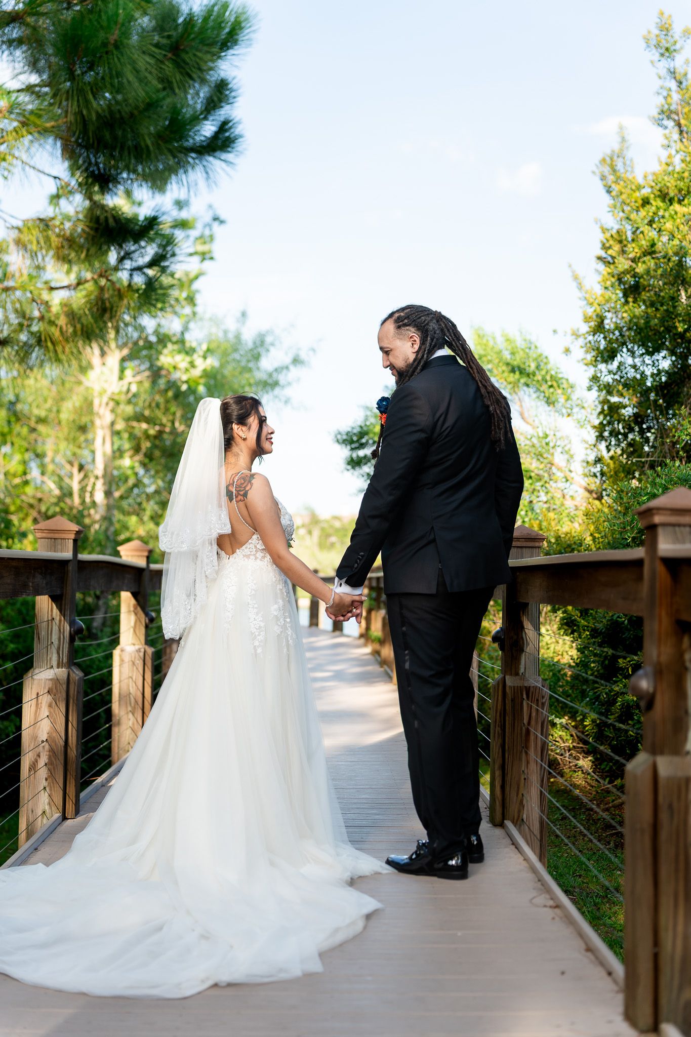 Bride and groom facing each other holding hands on scenic outdoor wooden bridge.