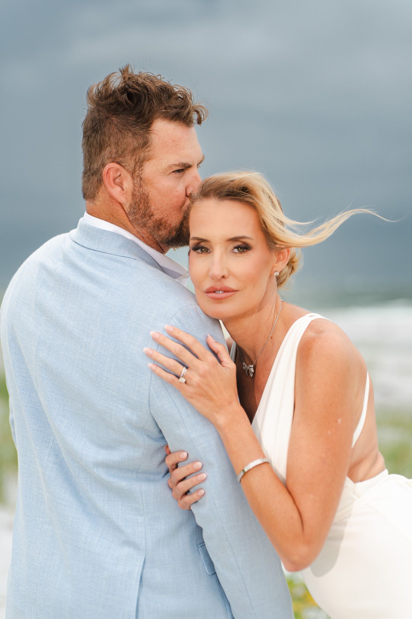 Groom in light blue suit tenderly kissing bride on cheek while she embraces him at beach wedding.