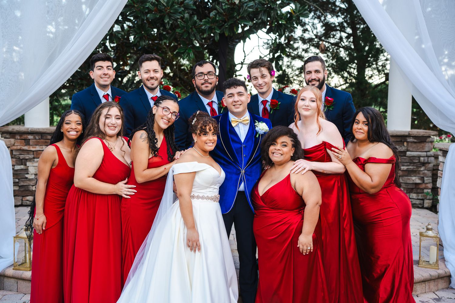 Wedding party poses together with bridesmaids in red dresses and groomsmen in navy suits at outdoor ceremony.