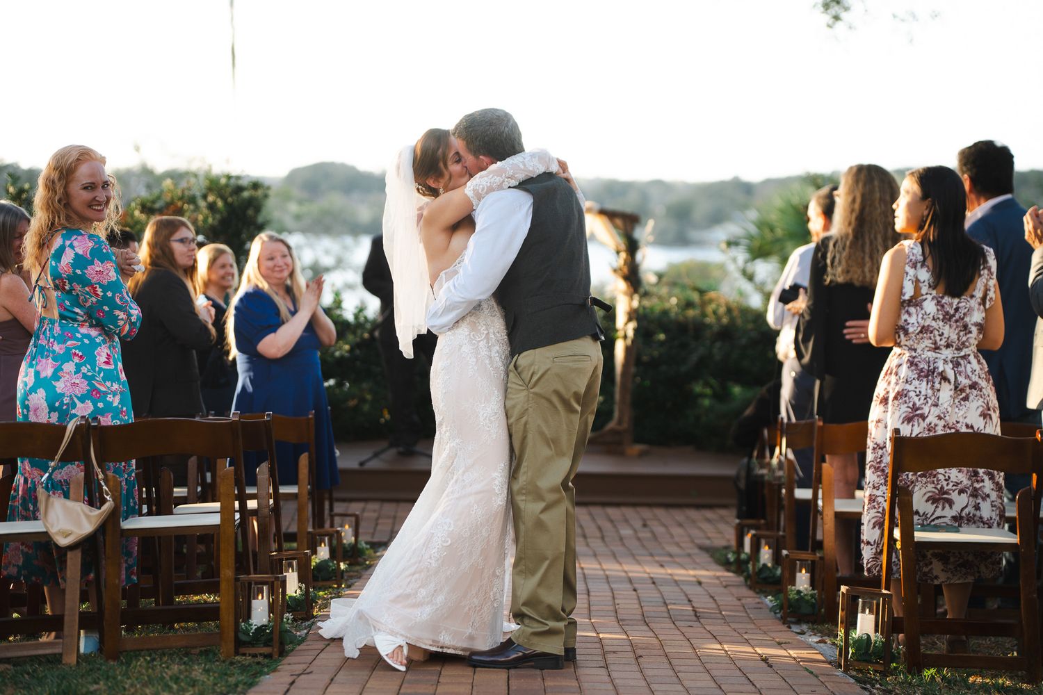 Bride and groom share romantic kiss in the aisle surrounded by wedding guests outdoors.