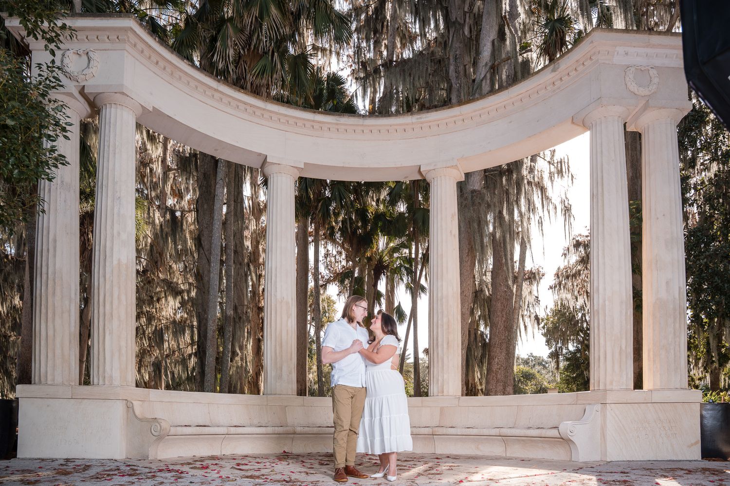 Couple embracing under a elegant white stone colonnade arch surrounded by tall trees.
