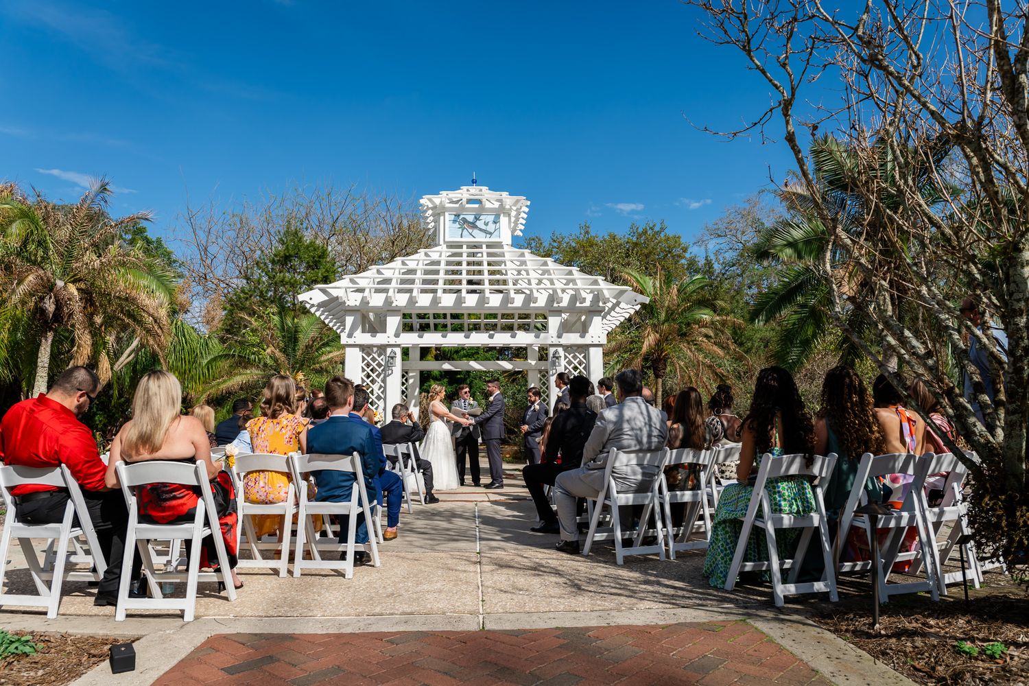 Outdoor wedding ceremony with guests seated in white chairs facing a white lattice gazebo surrounded by palm trees.