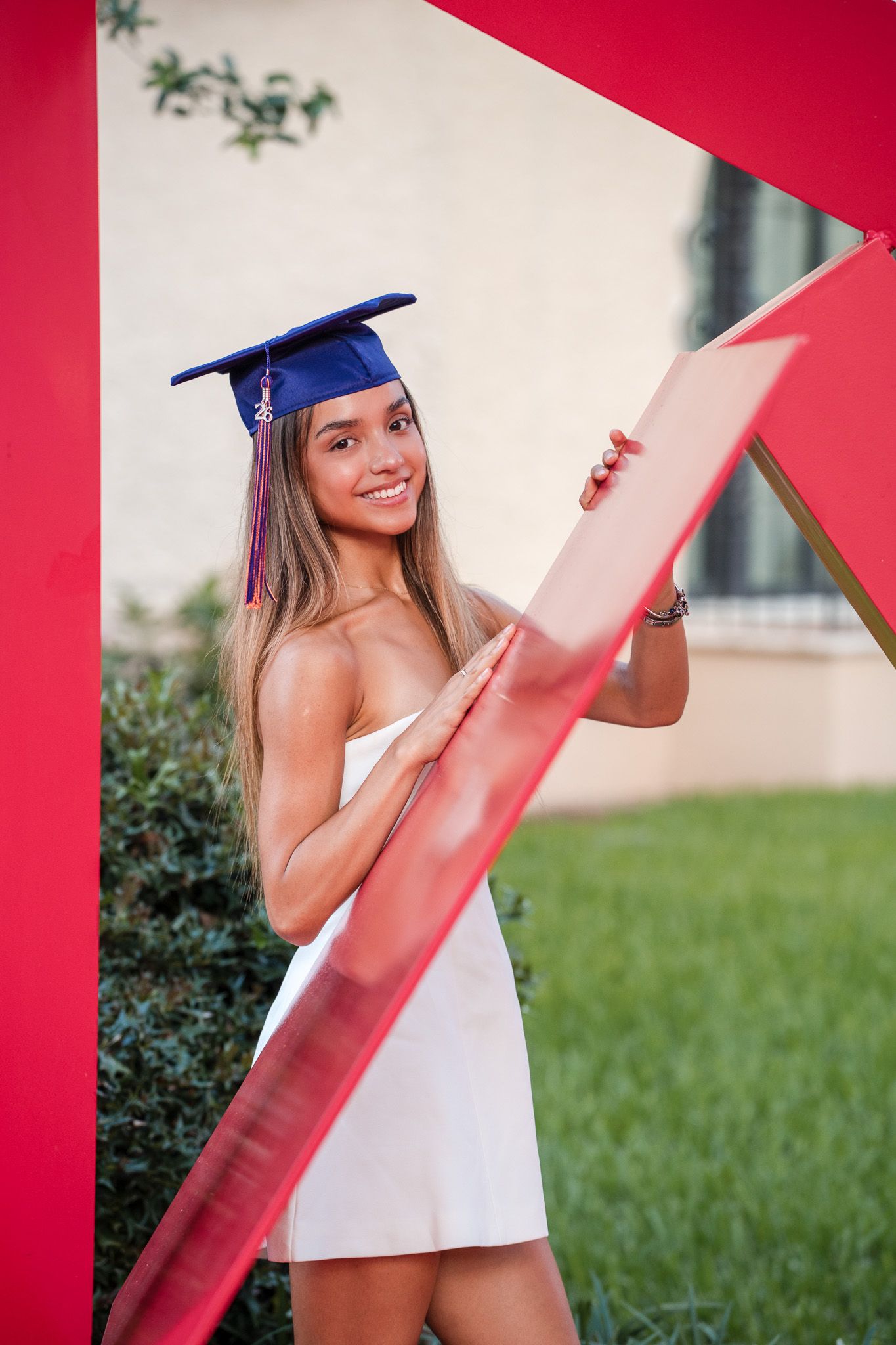 Smiling graduate in blue cap and white dress posing by red sculpture outdoors.