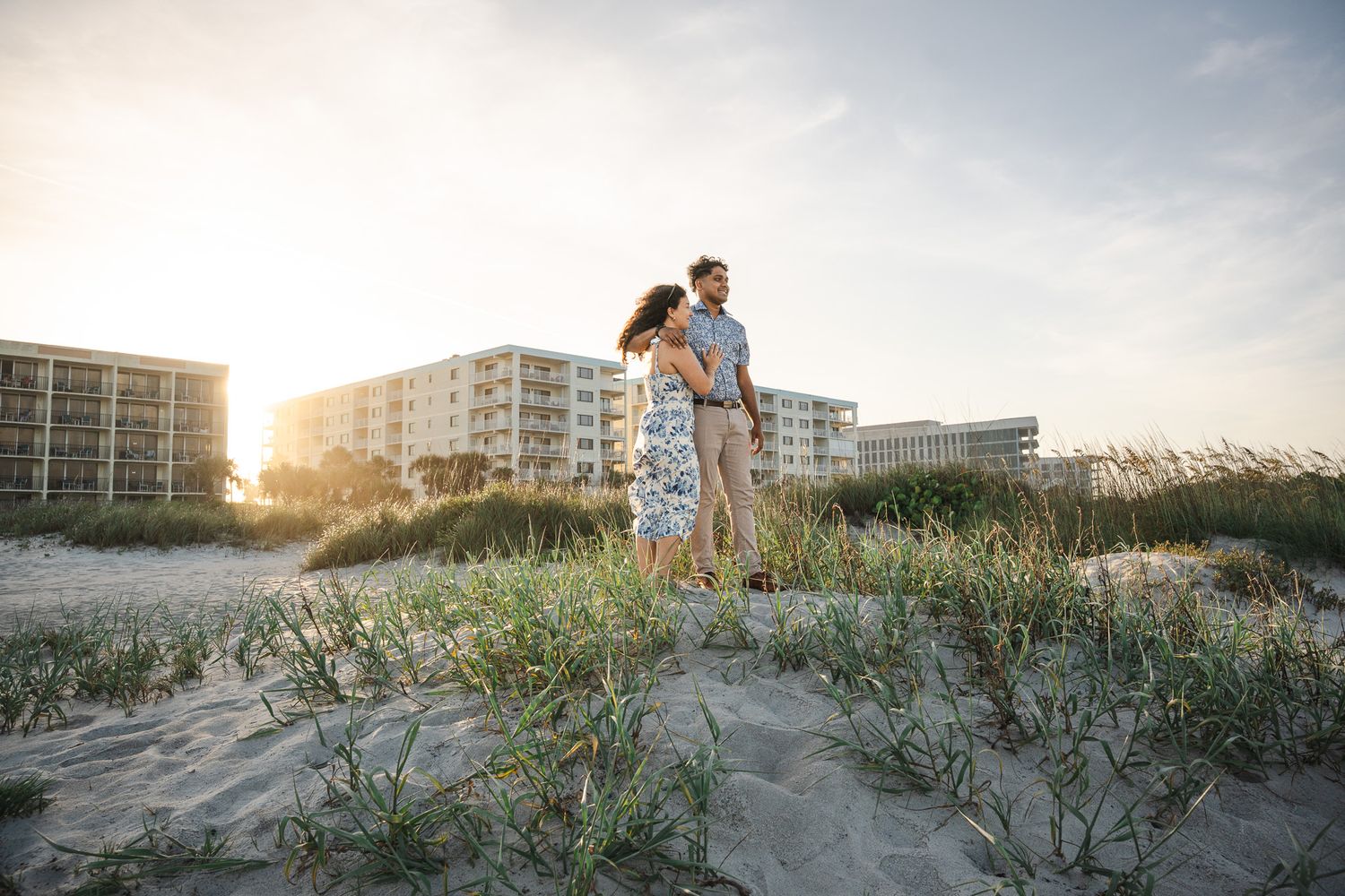 A couple stands together on a sandy beach at sunset with coastal buildings and sea grass in the background.