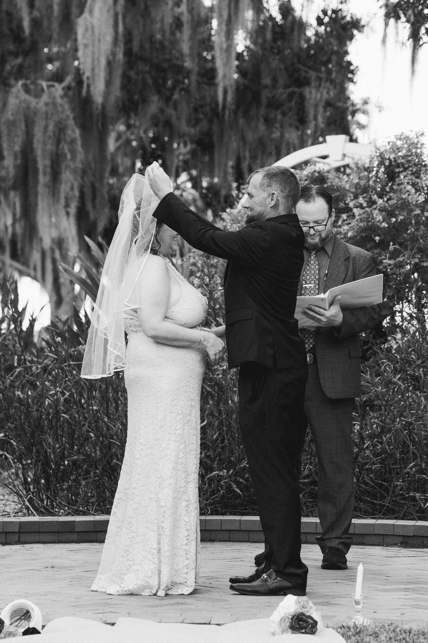 Groom lifts bride's veil during outdoor wedding ceremony while officiant reads vows, surrounded by Spanish moss trees.