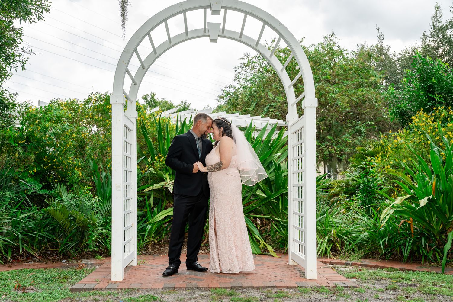 Bride and groom share a romantic moment under a white arch surrounded by lush greenery at an outdoor wedding.