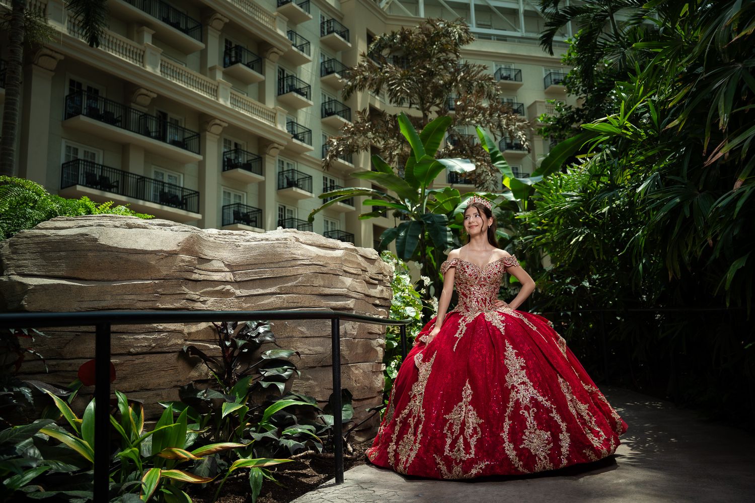 Young woman in red and gold ball gown quinceañera dress poses in lush hotel atrium with tropical plants.