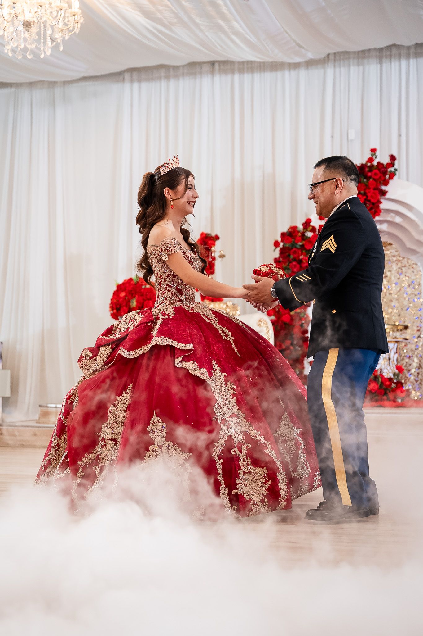 Girl in red quinceañera gown dances with man in military uniform on foggy dance floor at elegant party.