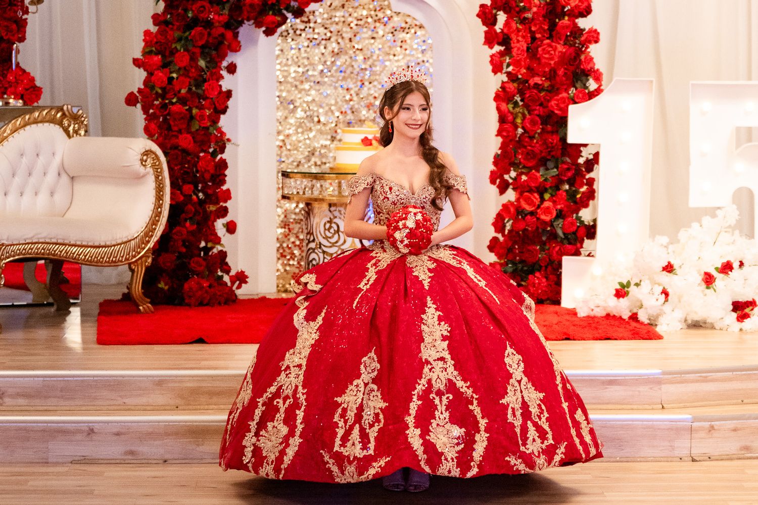 Young woman in red and gold quinceañera ball gown poses elegantly before floral rose backdrop.