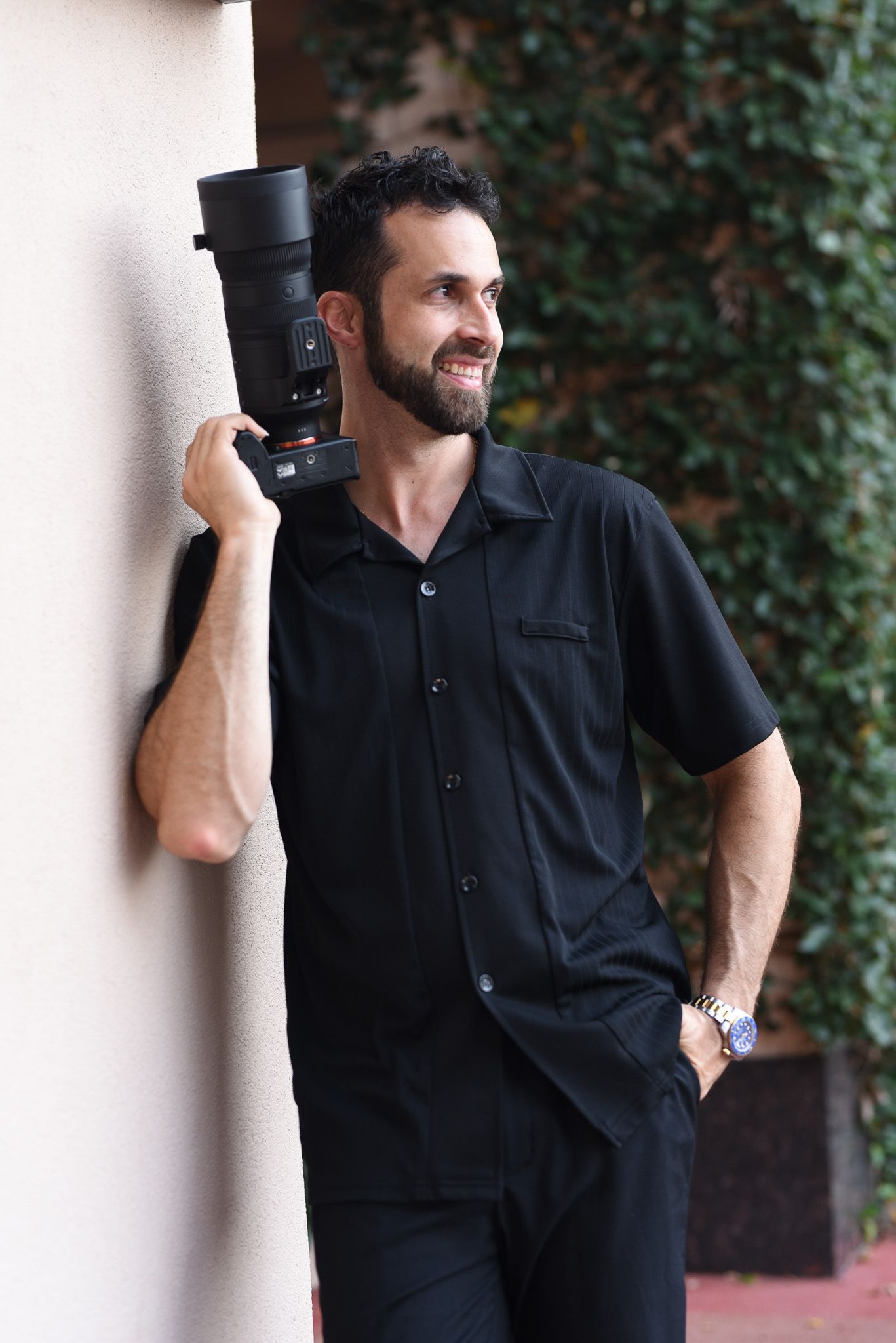 Smiling bearded man in black shirt holding a professional camera with telephoto lens against a wall outdoors.