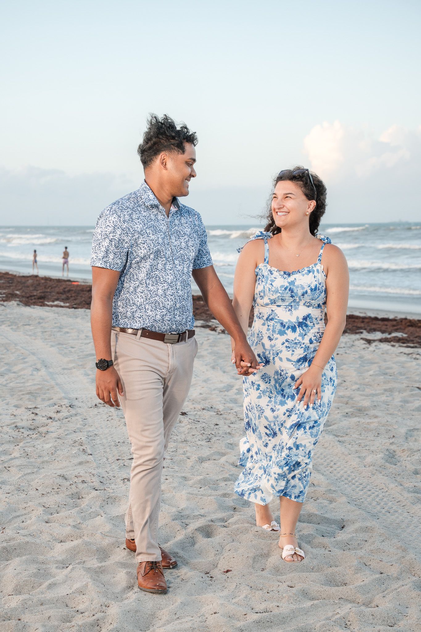 A couple walks hand-in-hand on a sandy beach, wearing matching blue floral outfits, smiling at each other with ocean waves behind.