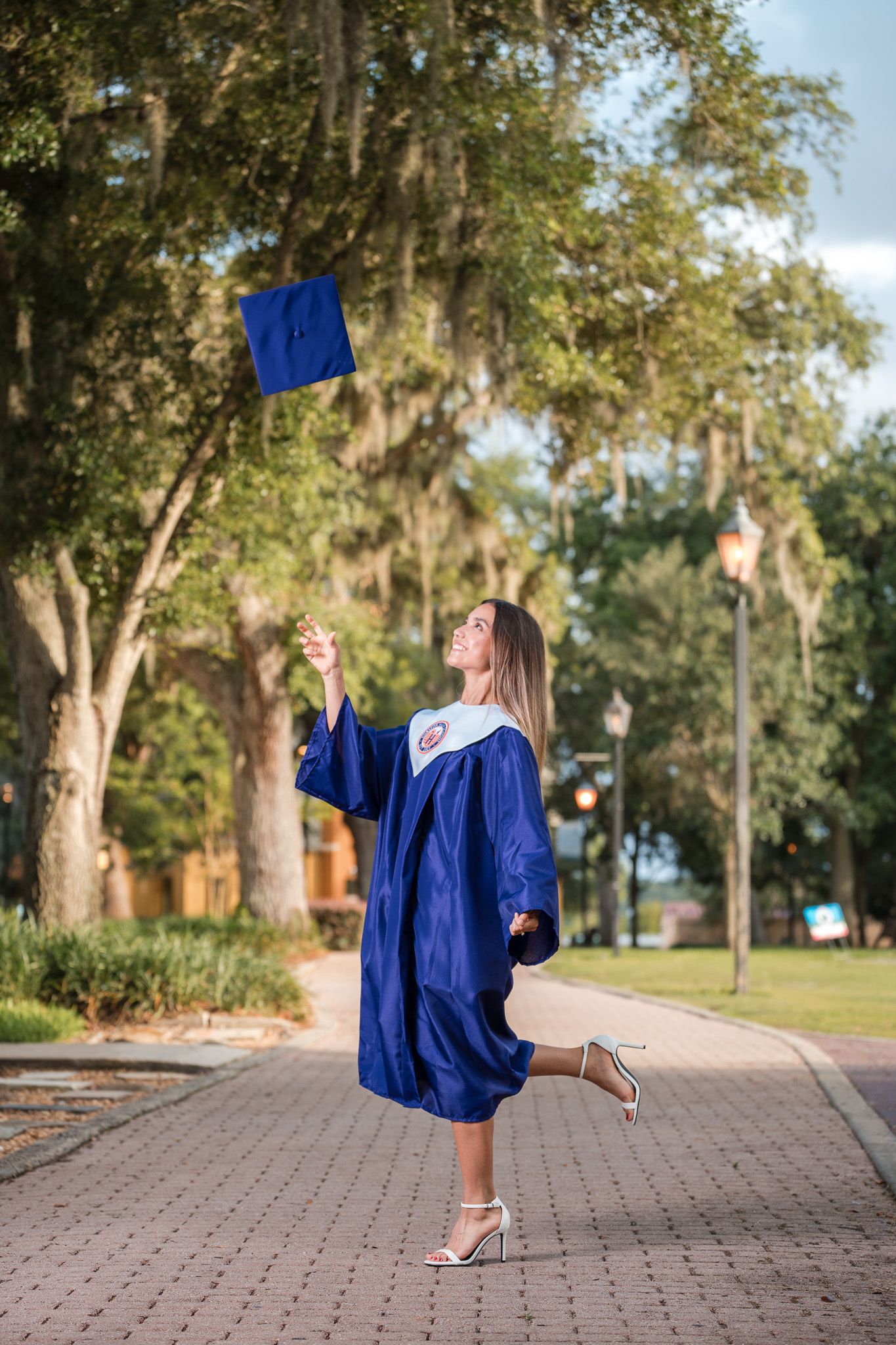 A young woman in a blue graduation gown tosses her cap in the air on a tree-lined campus walkway.