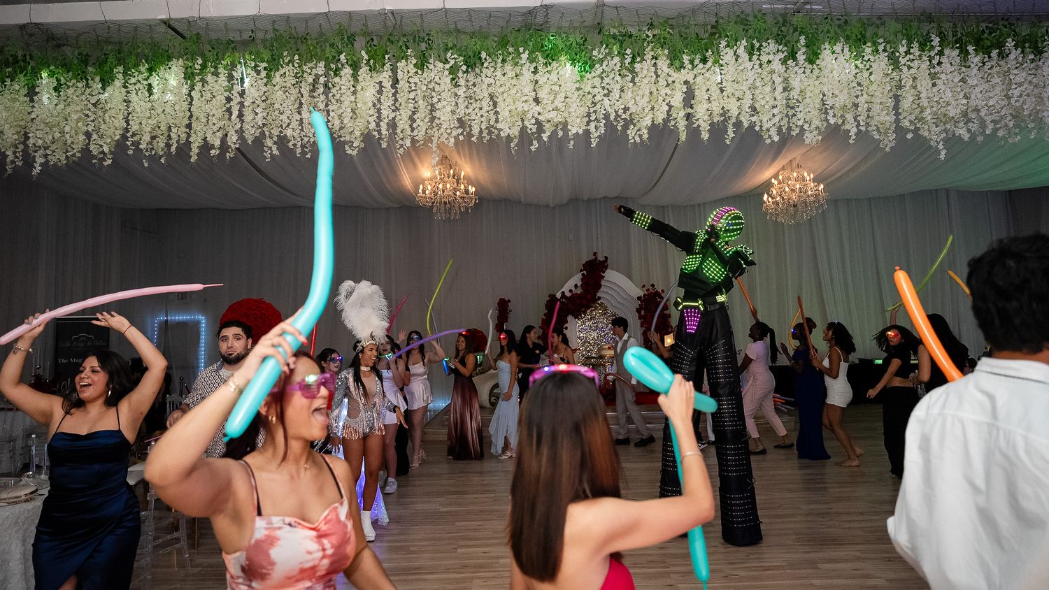 Guests dance at a festive party with balloon sticks, floral ceiling decor, and colorful lighting in an elegant venue.