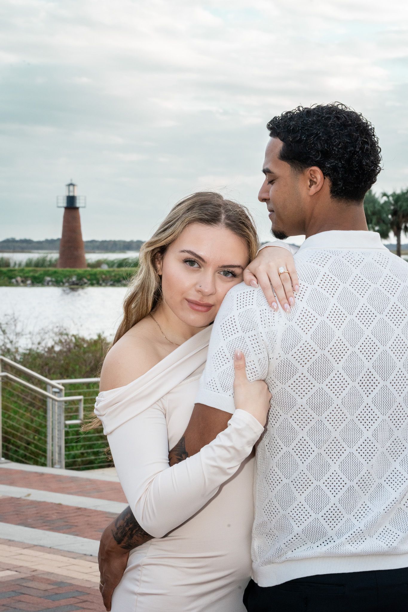 Woman in cream off-shoulder dress embracing man near waterfront with lighthouse backdrop.
