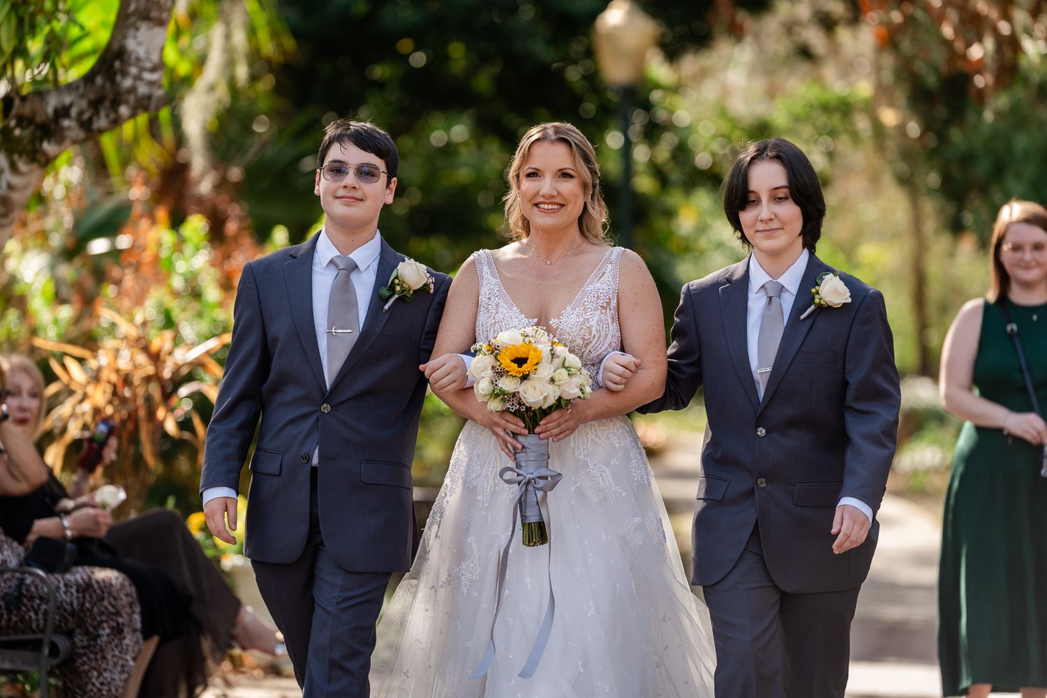 Bride walks down outdoor aisle with two groomsmen holding sunflower bouquet at garden wedding ceremony.