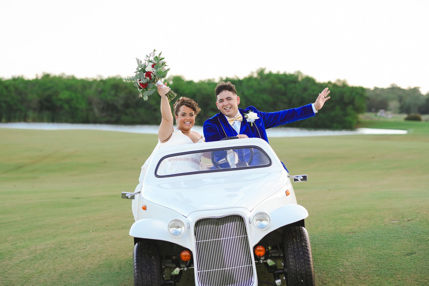 Bride and groom celebrate joyfully riding vintage white golf cart across scenic green golf course.