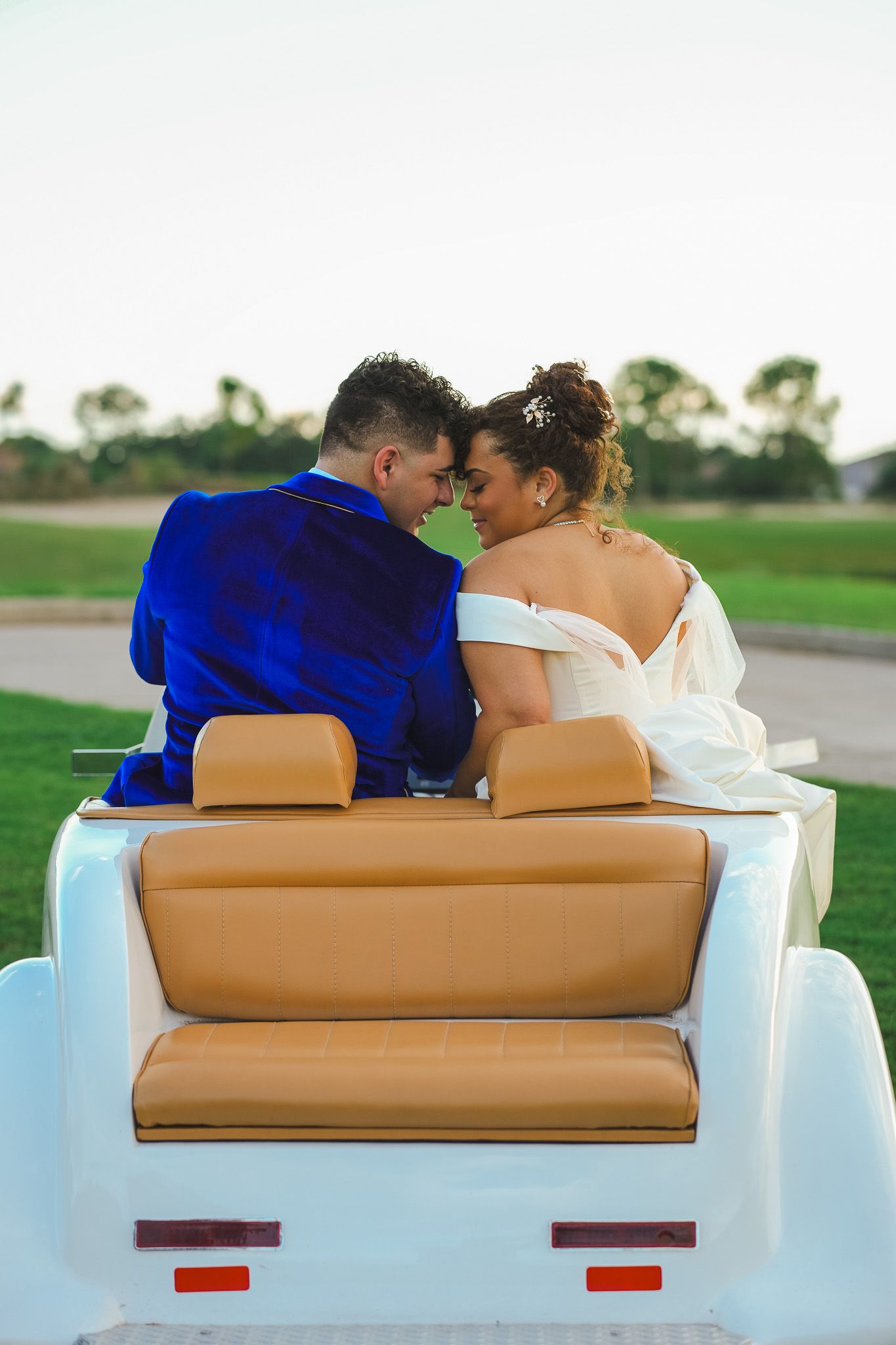 Bride and groom sitting close together in a vintage white golf cart on a green golf course.