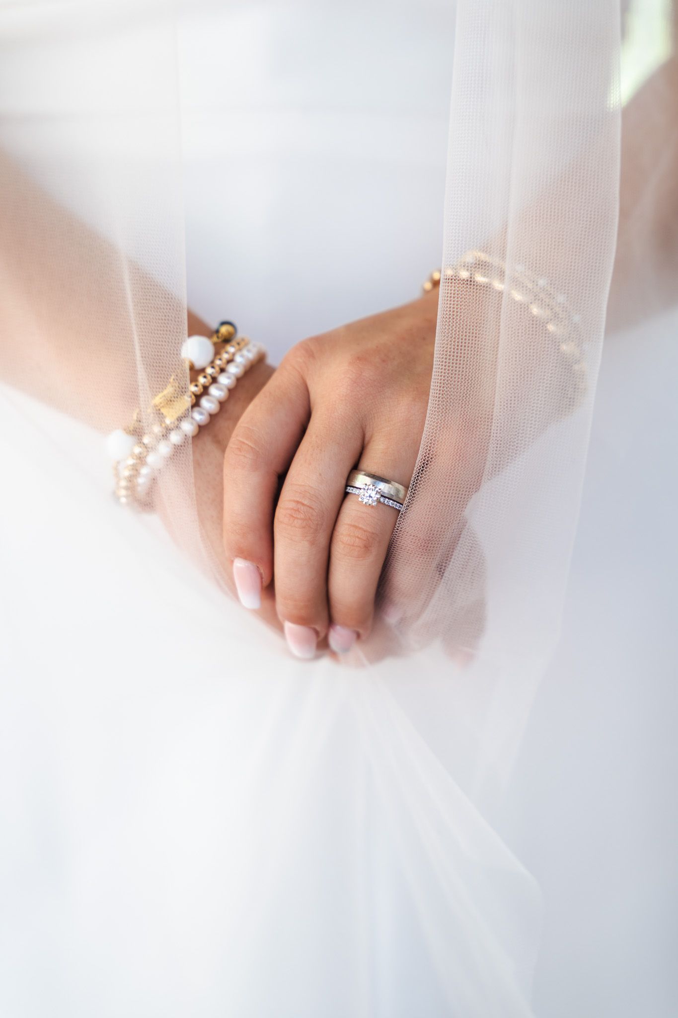 Close-up of bride's hands showing diamond wedding ring and pearl bracelets against white bridal gown.