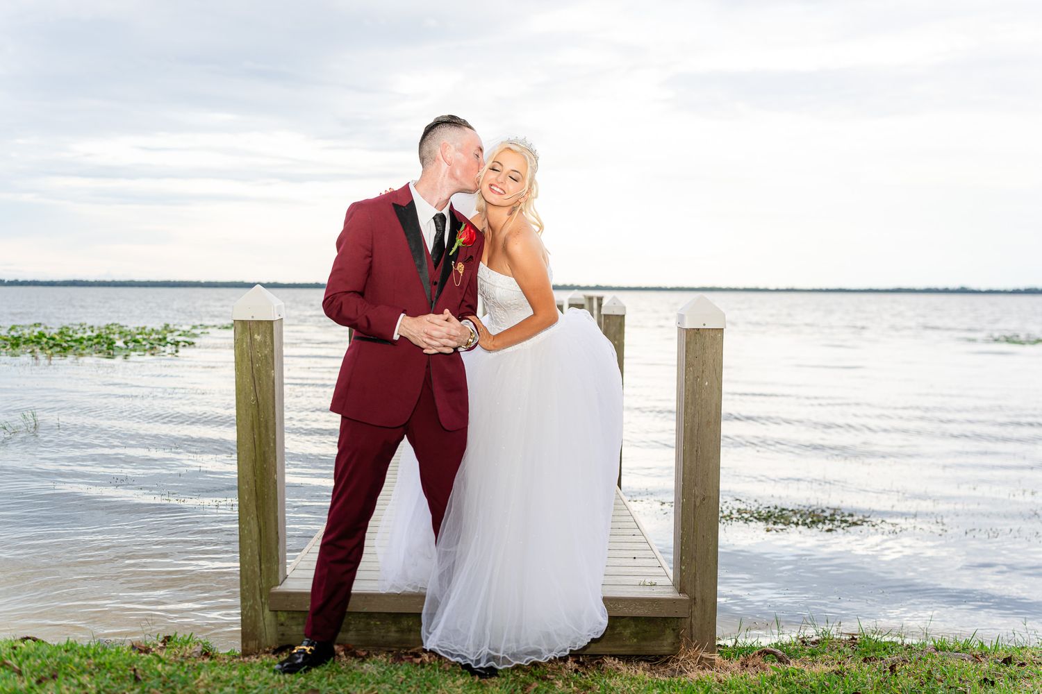Bride and groom posing on a wooden dock by a lake, groom kissing bride on her cheek.