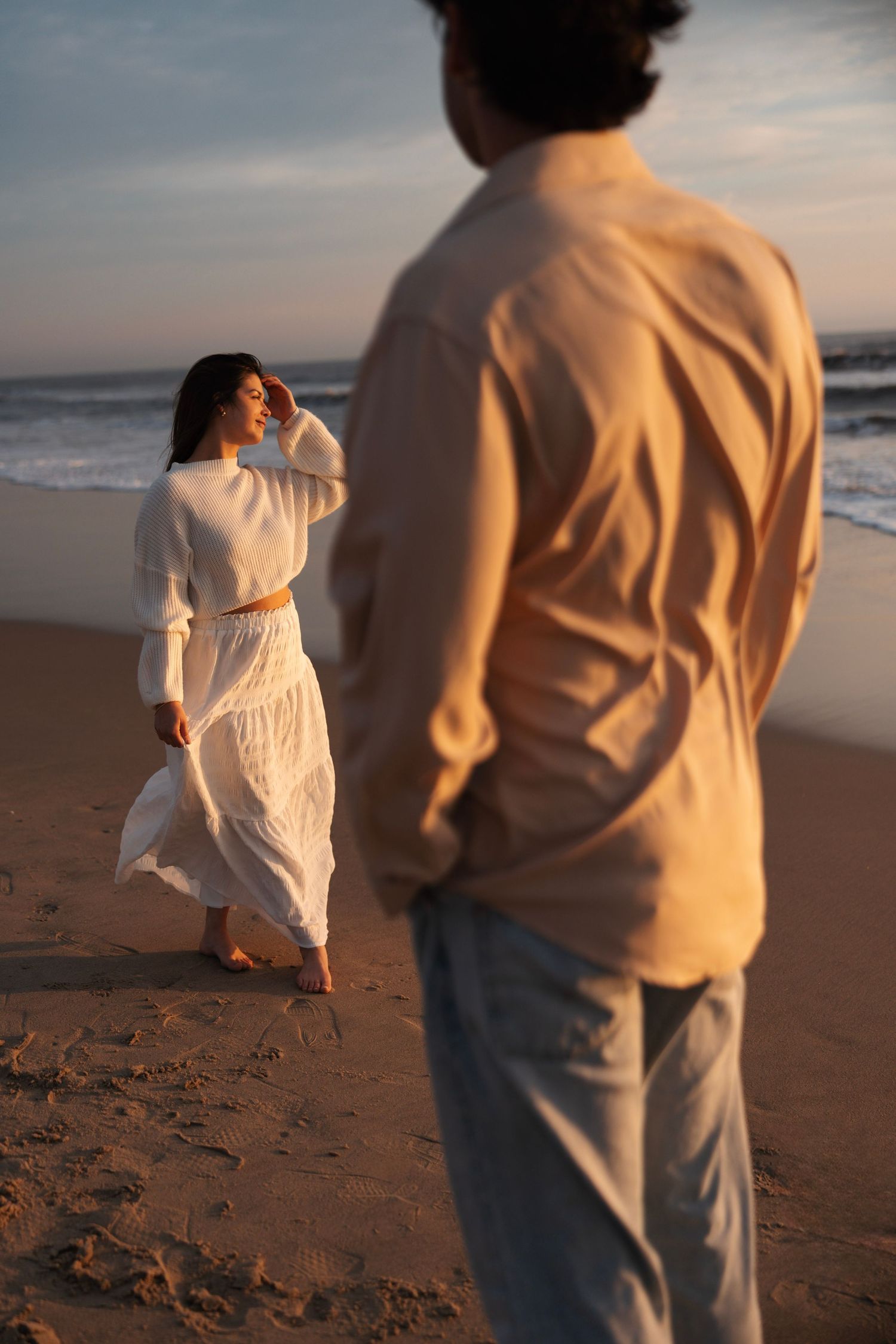 Woman in flowing white dress poses on beach at golden hour while man watches in foreground.