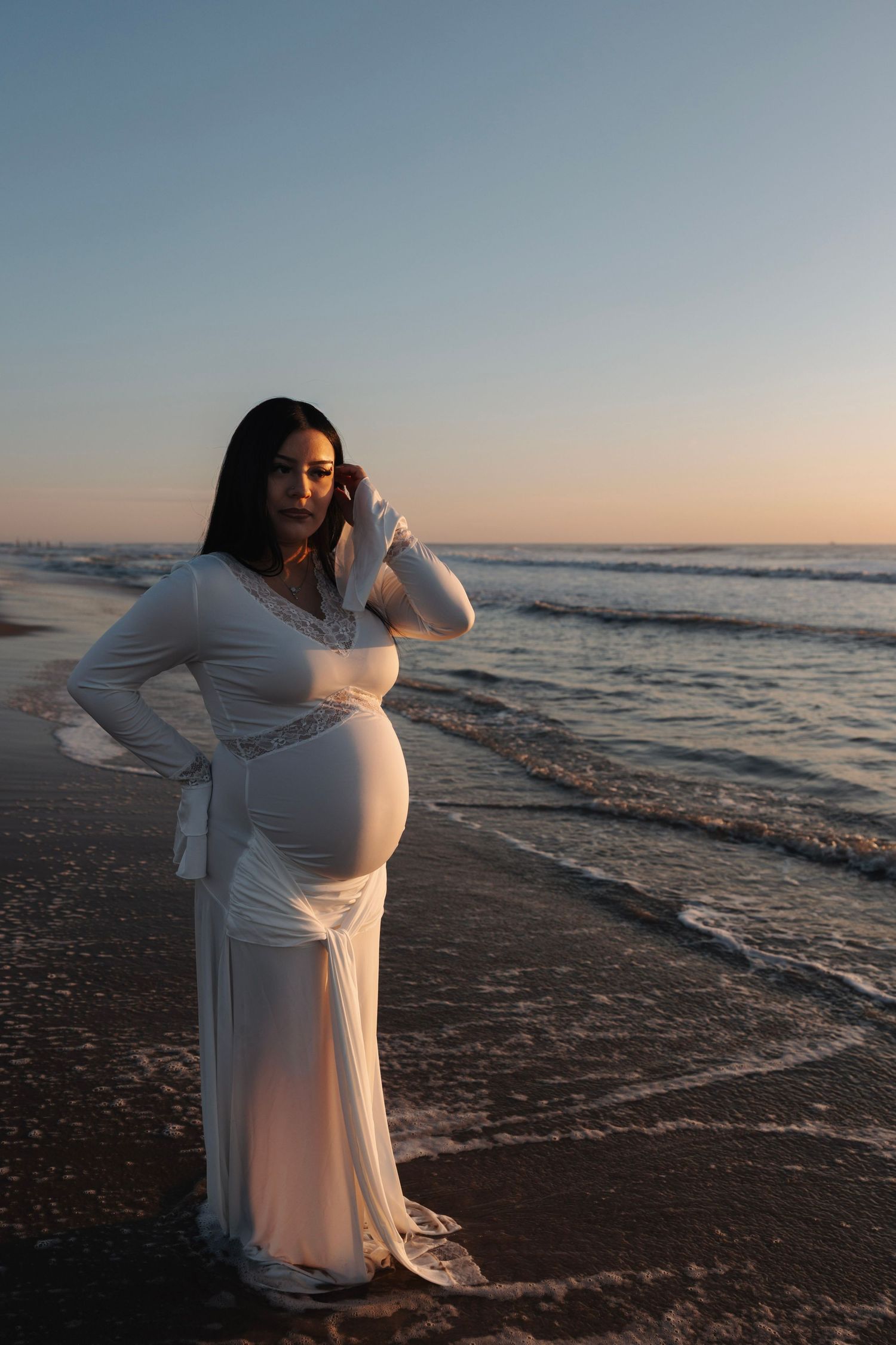 Pregnant woman in white gown posing gracefully on beach at sunset.