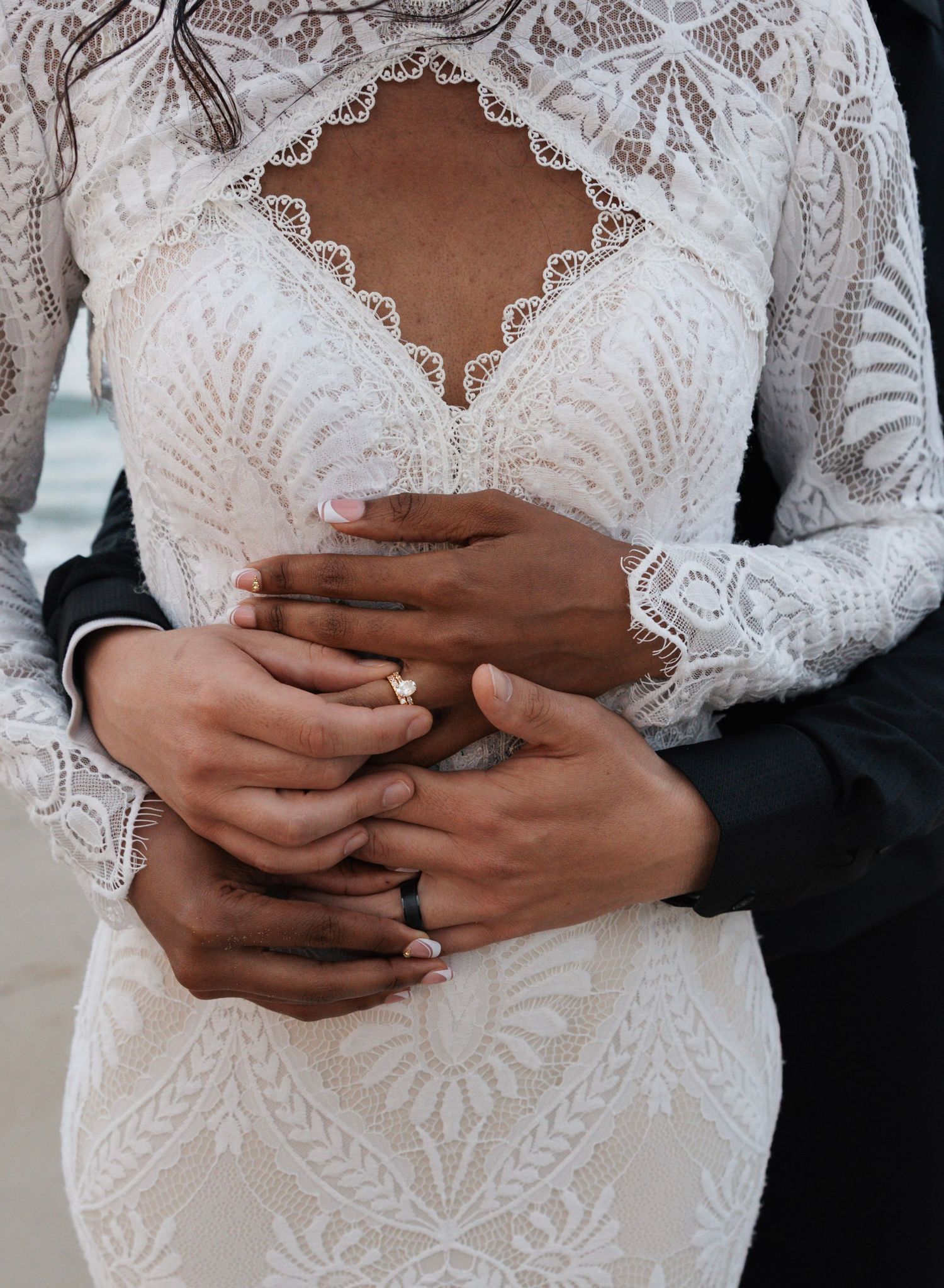 Couple holding hands showing wedding rings, bride wearing lace long-sleeve gown on beach.