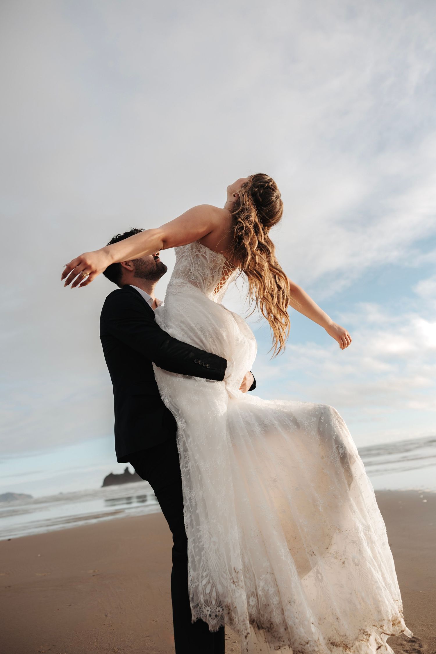 Groom lifting bride in white lace wedding dress on sandy beach with ocean waves in background.
