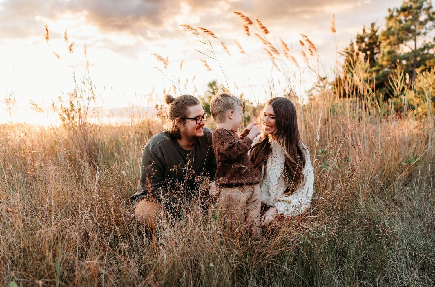 Happy family with child sitting together in golden sunset field of tall grass at dusk.