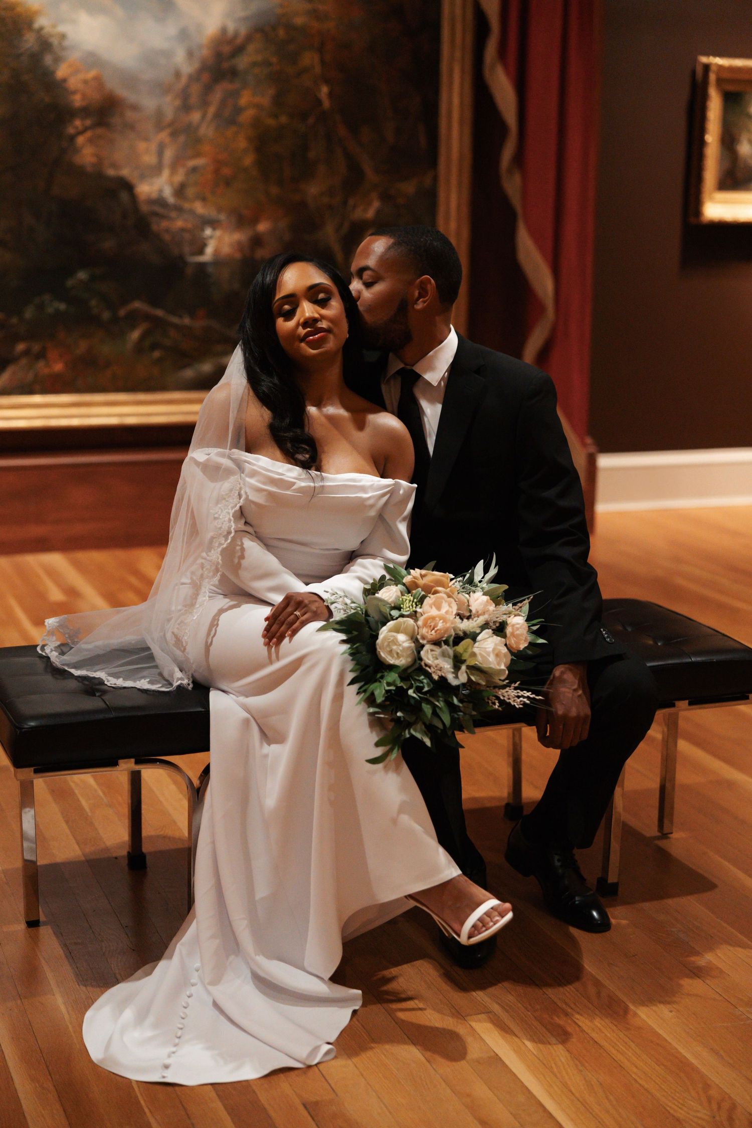 Groom kissing bride on cheek while seated in art museum holding elegant bridal bouquet.