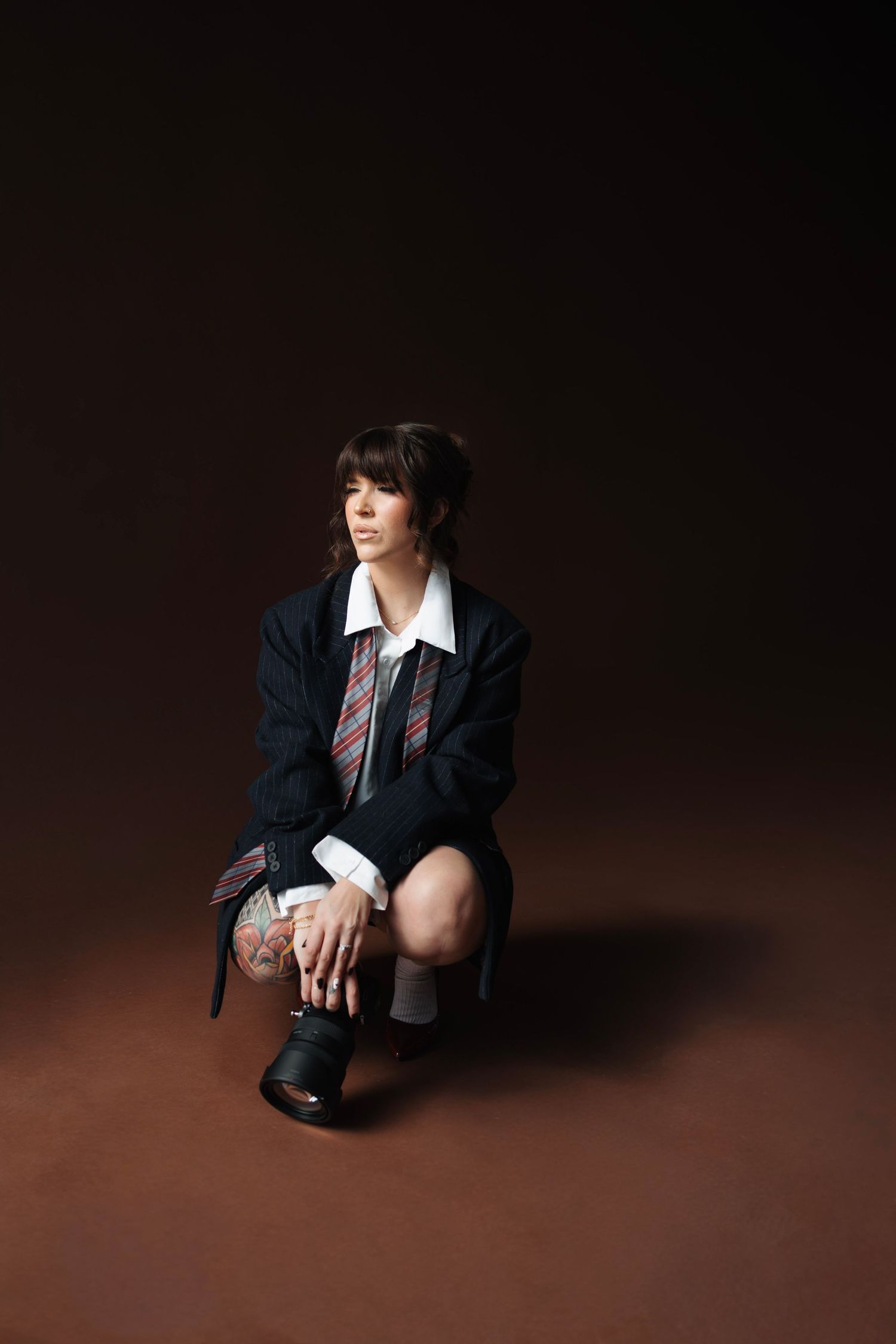 Photographer crouching with camera lens, wearing blazer and tie against dark brown studio background.