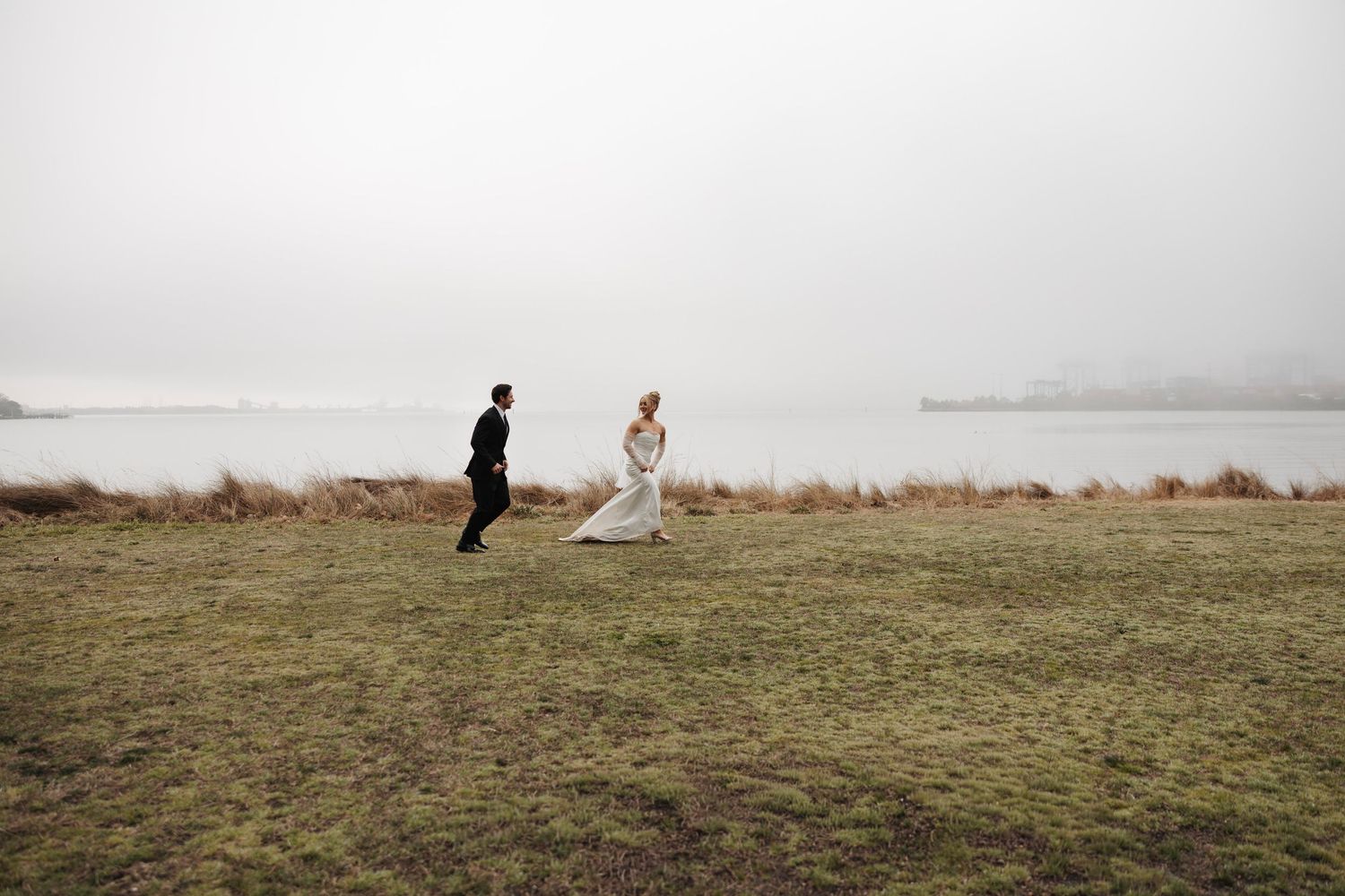 Bride and groom running through misty green field near lake on their wedding day.