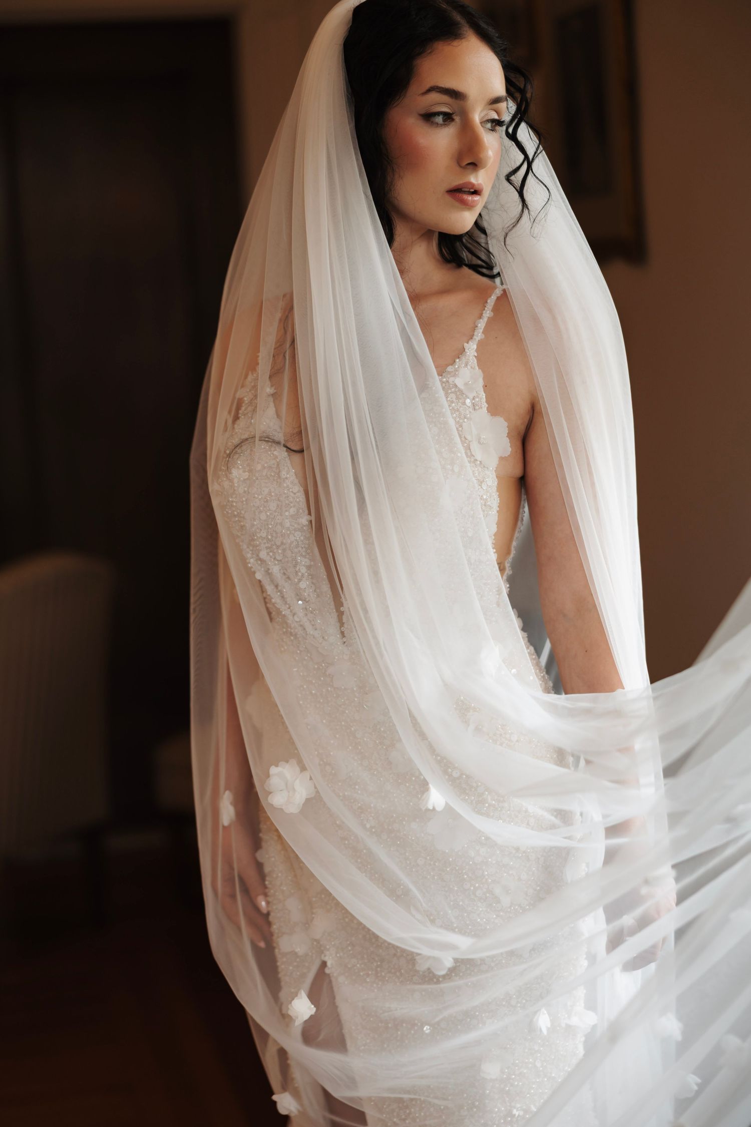 Beautiful dark-haired bride in lace wedding dress holding flowing white veil in soft indoor light.
