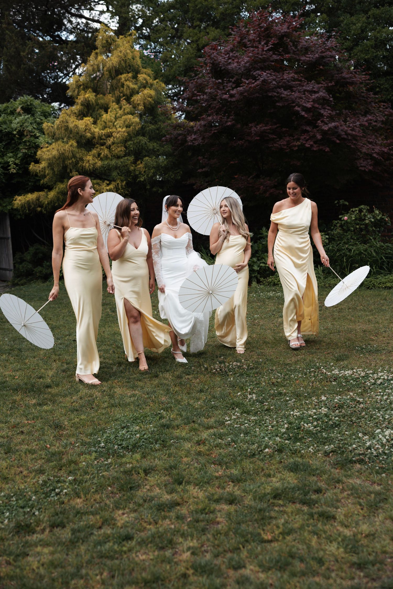 Bride and bridesmaids in yellow gowns holding white parasols walking on garden lawn.
