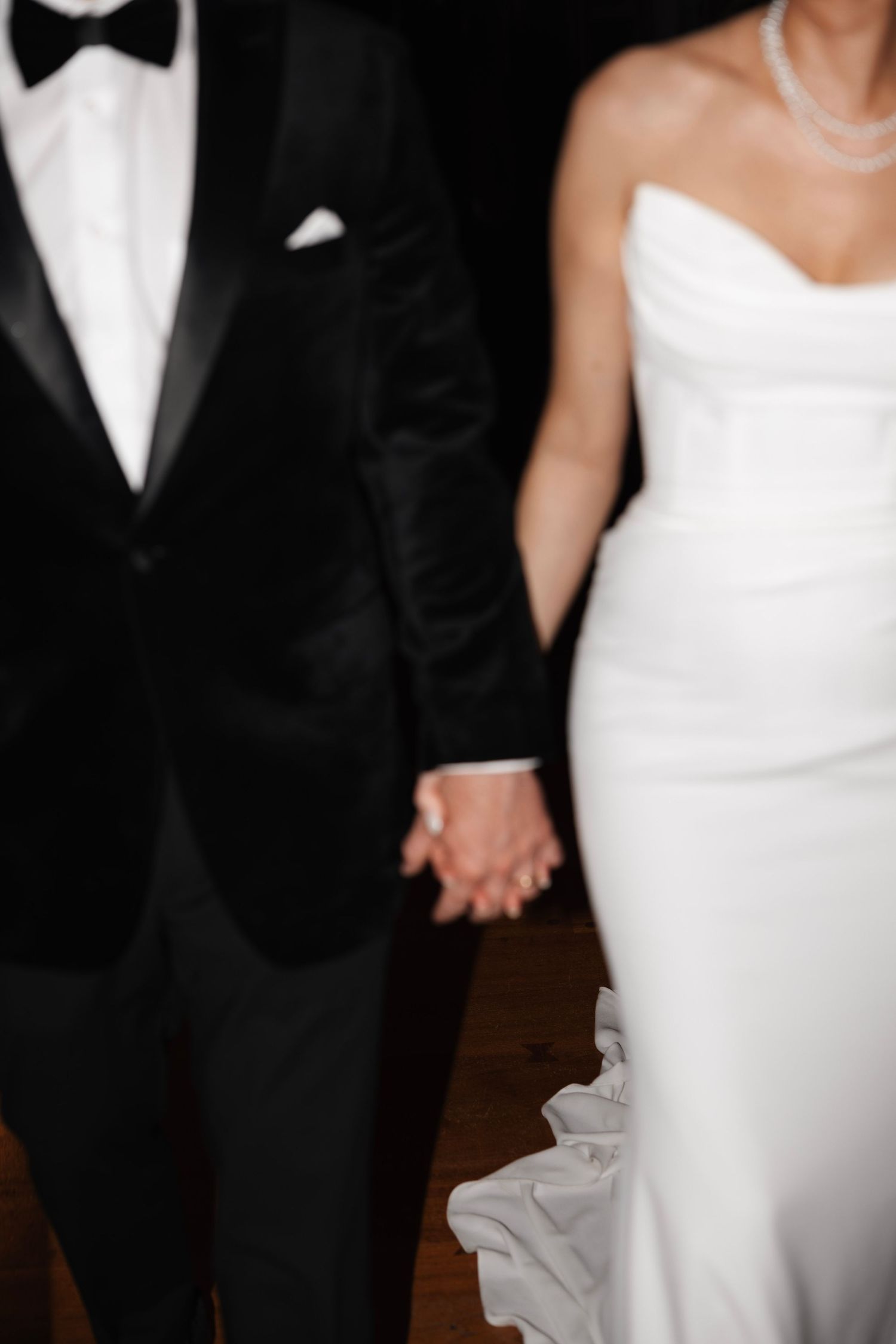 Bride in strapless white gown and groom in black tuxedo holding hands.