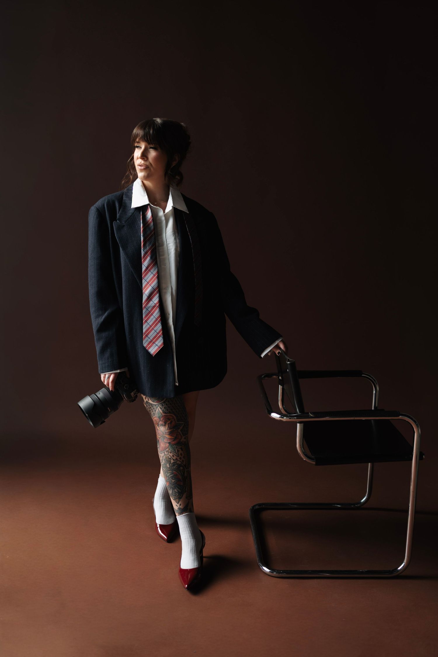 Woman in oversized blazer and striped tie holding camera beside modern chair with tattooed legs and red heels.