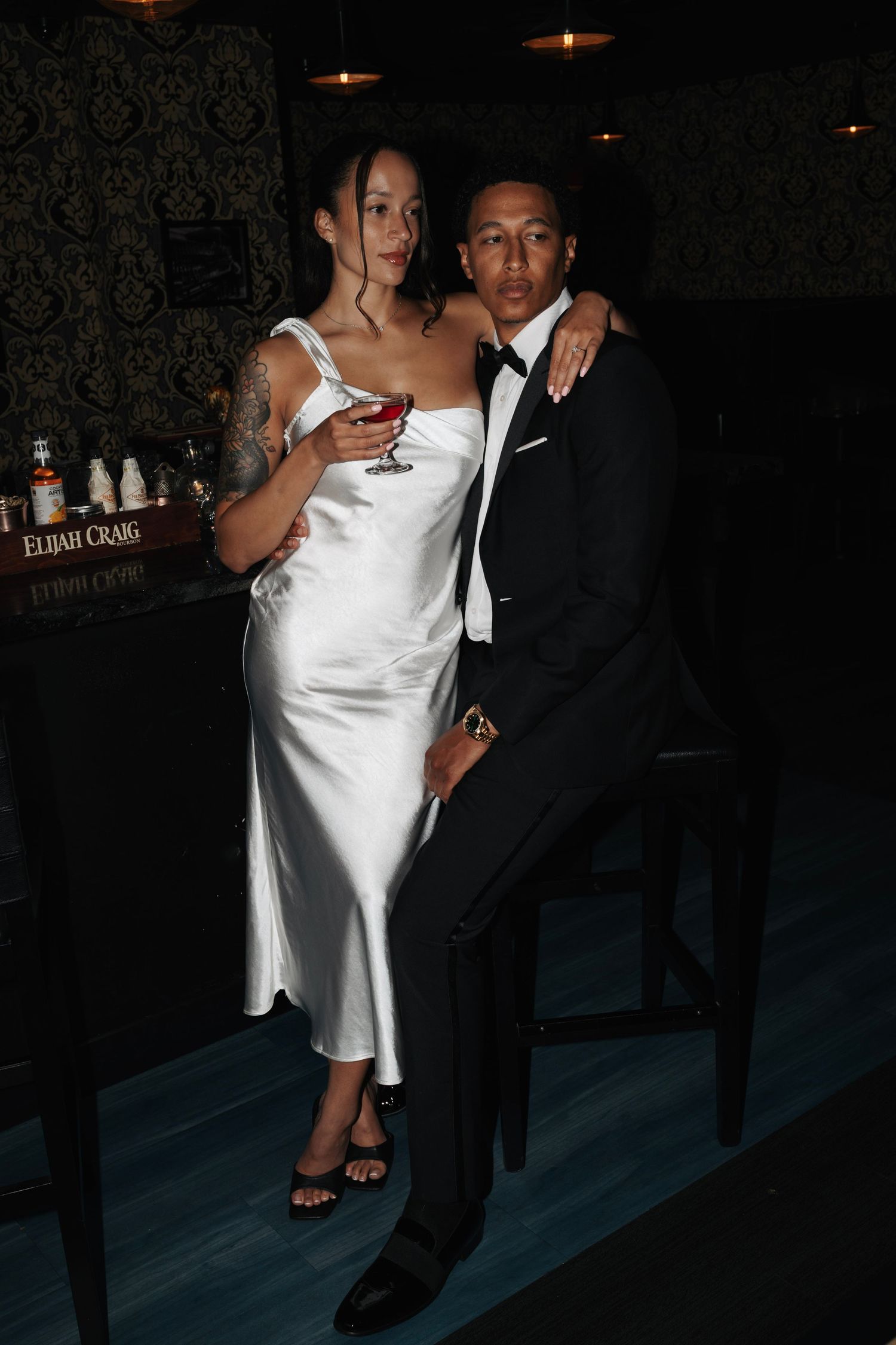 Couple posing at bar, woman in white satin slip dress holding wine glass, man in black tuxedo.
