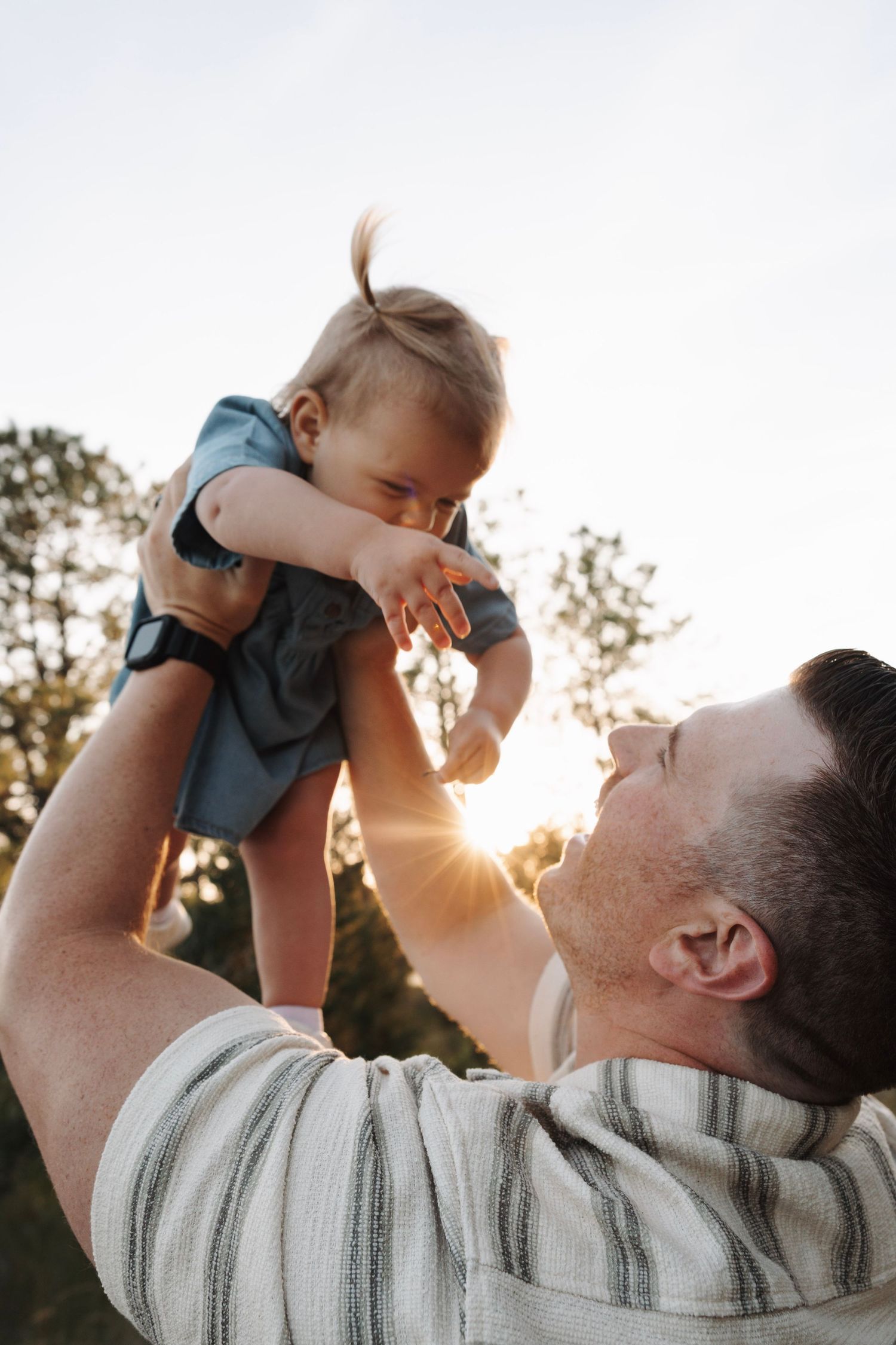 Father lifting smiling baby in the air outdoors with golden sunlight glowing through trees behind them.