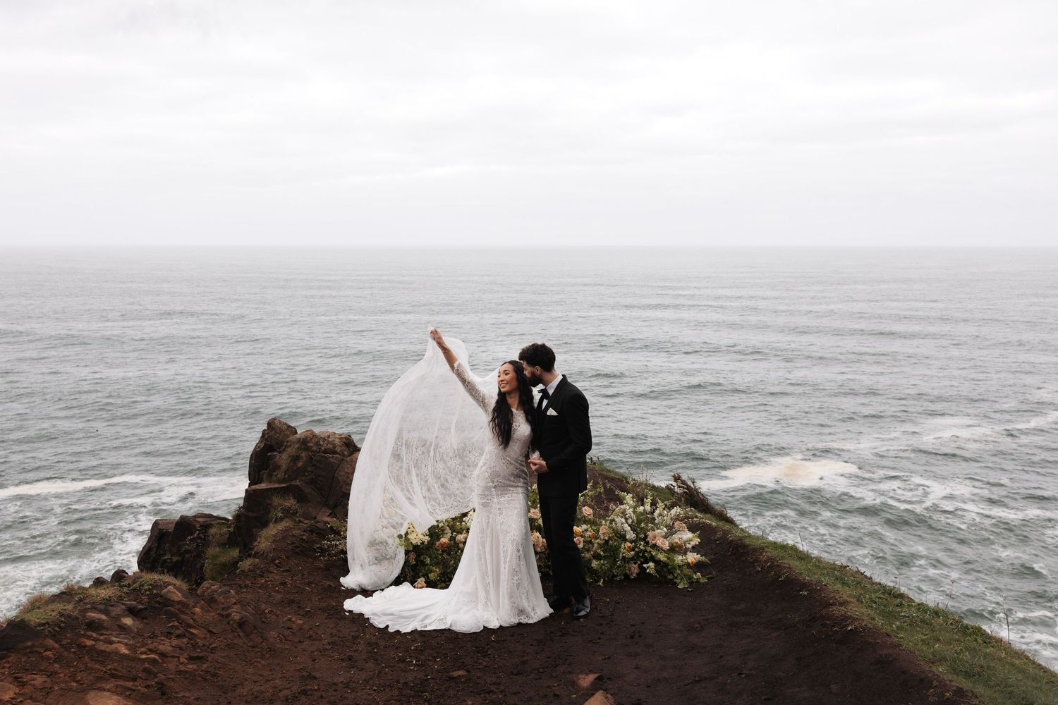 Bride and groom kiss on dramatic coastal cliff edge with veil flowing in wind over ocean backdrop.