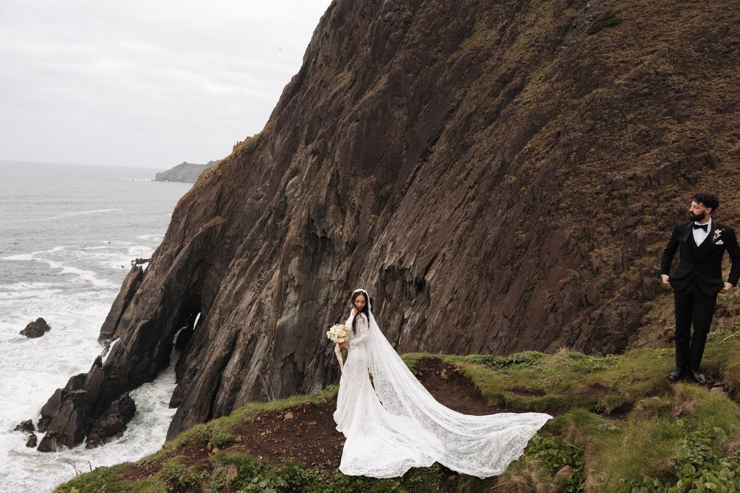 Dramatic wedding portrait of bride and groom on rugged coastal cliffs with ocean waves below.