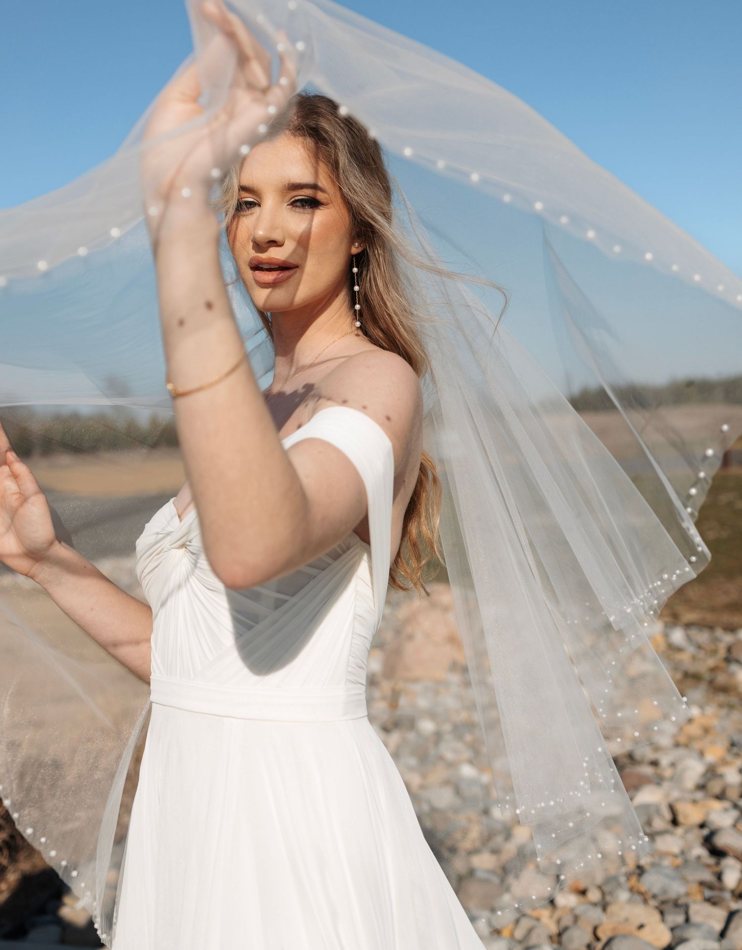 Blonde bride in off-shoulder white gown tosses pearl-edged veil against a bright blue sky outdoors.