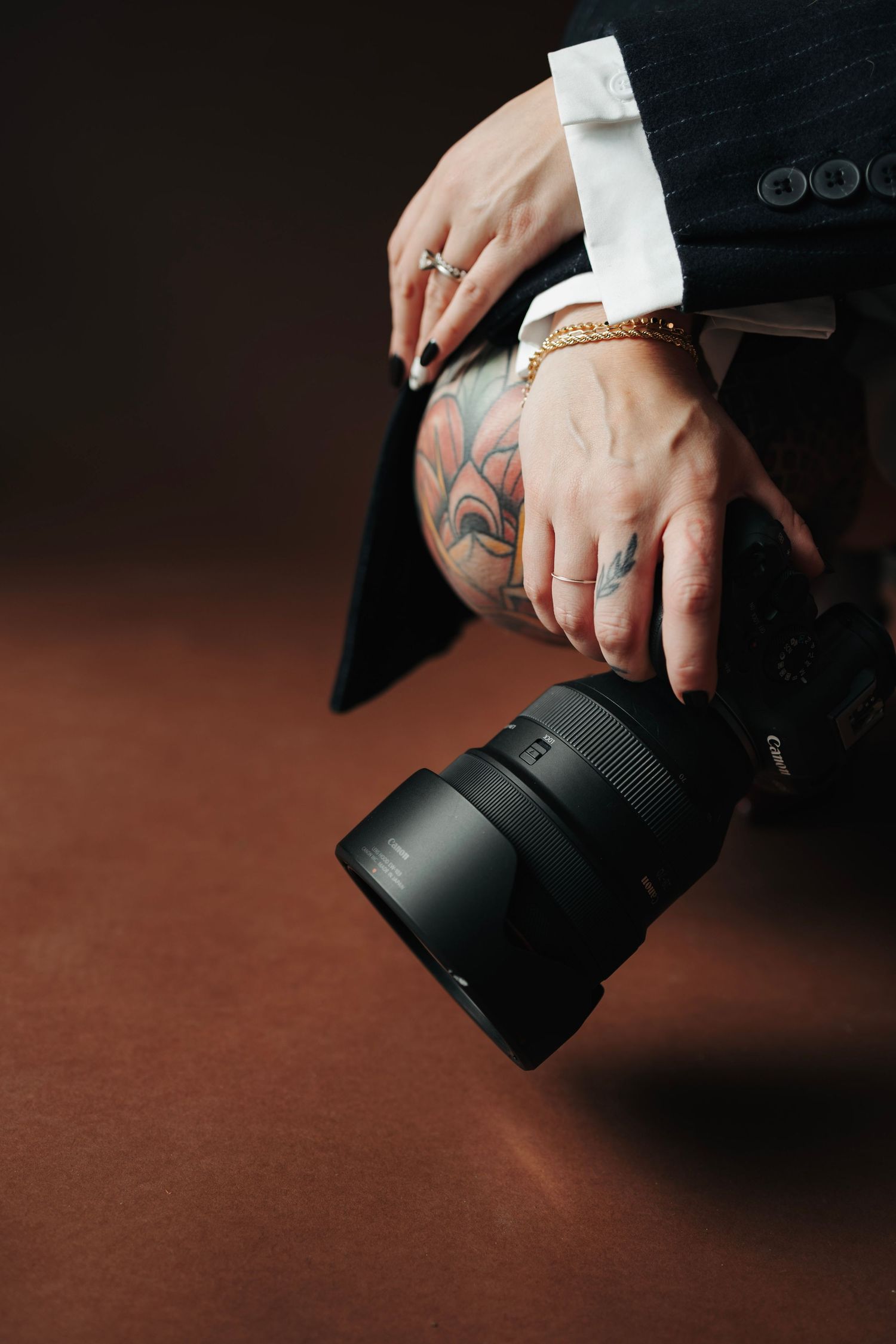 Tattooed hands holding professional DSLR camera with large lens in dark studio setting.