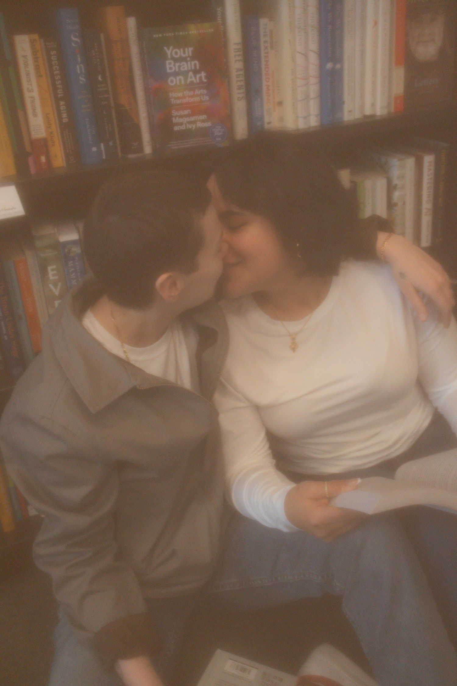 Two people sharing a kiss while sitting in front of a bookshelf filled with colorful books.