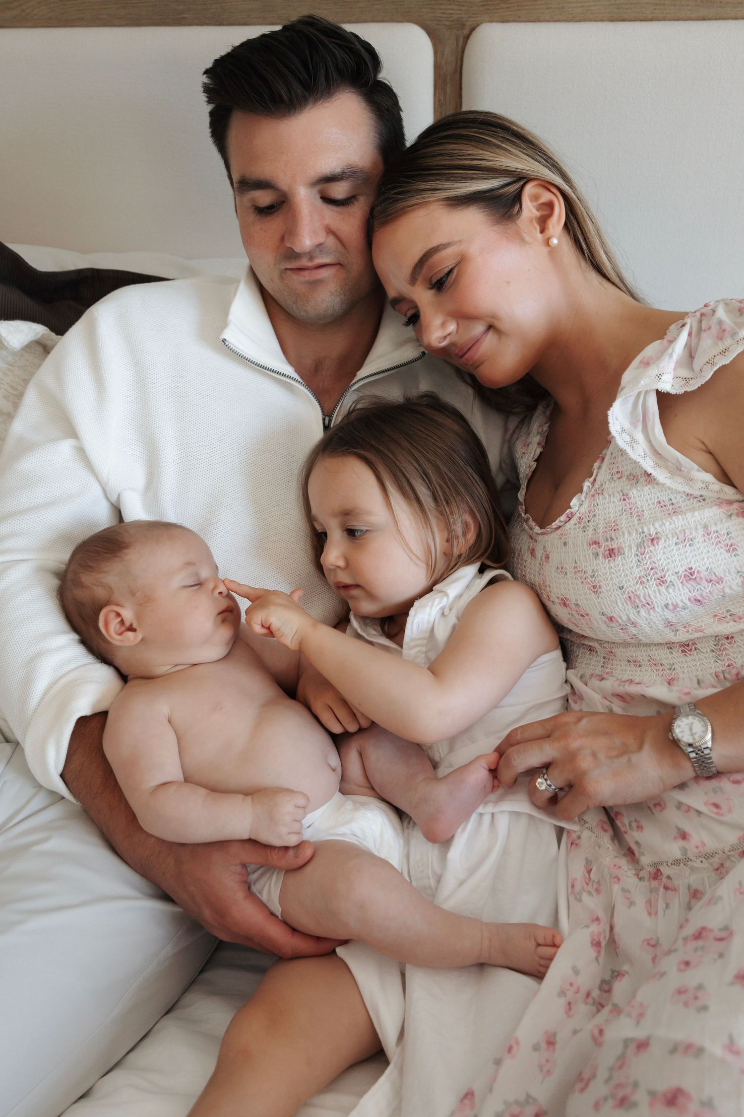 Happy family with father, mother, toddler, and newborn baby cuddling together on a white bed.