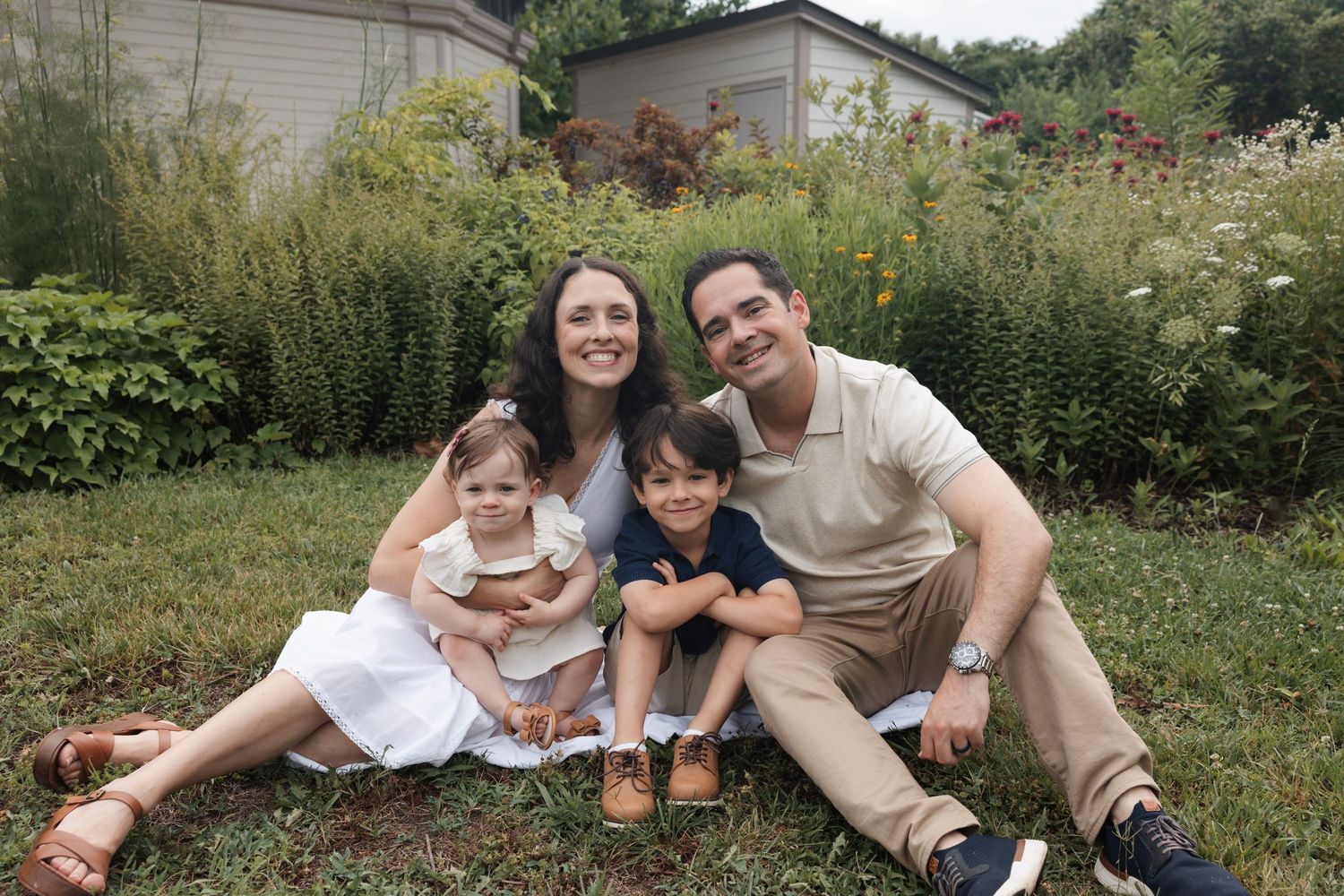 Happy family of four smiling together while sitting on grass in lush green backyard garden.