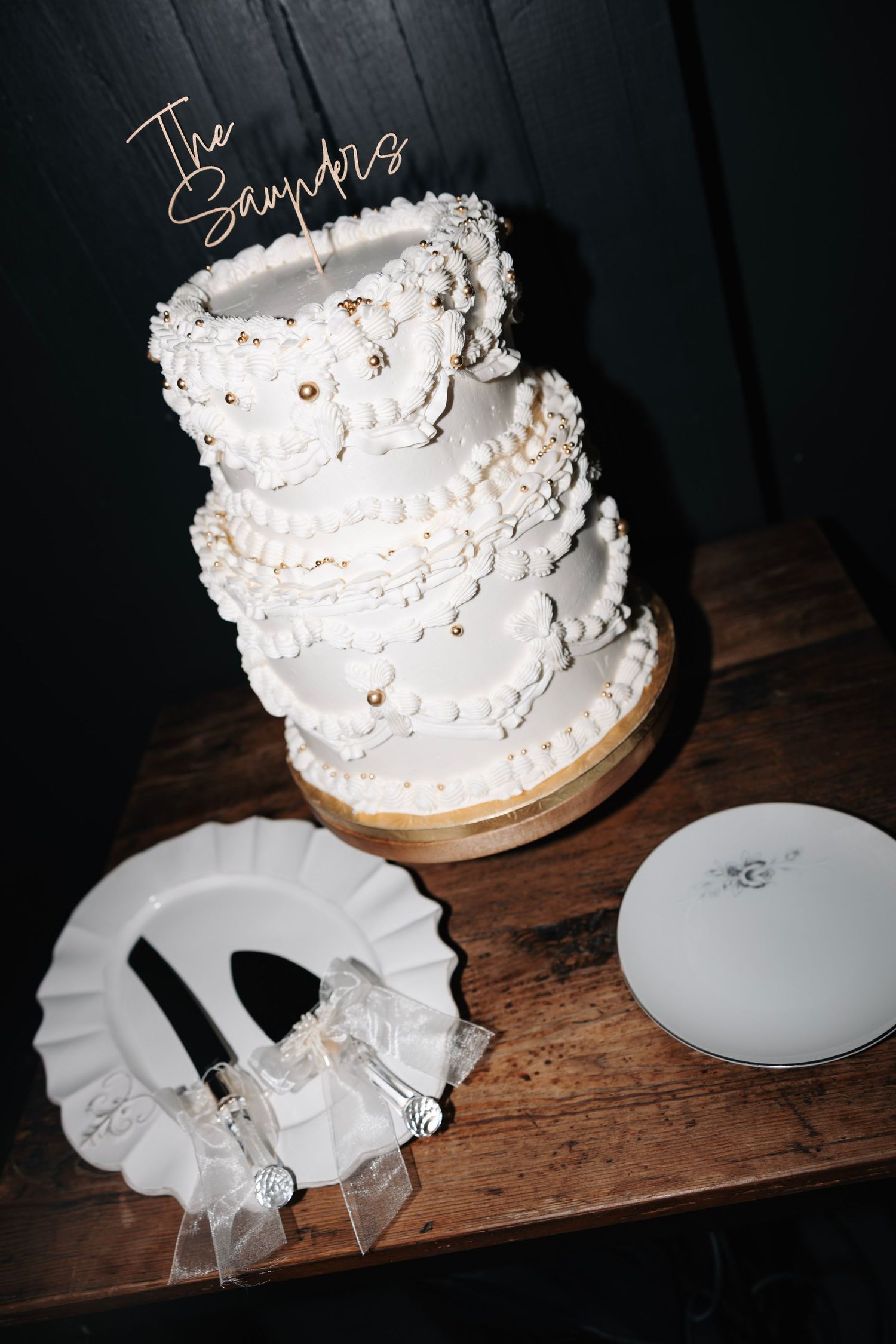 Three-tier white wedding cake with floral details and custom name topper on wood table.
