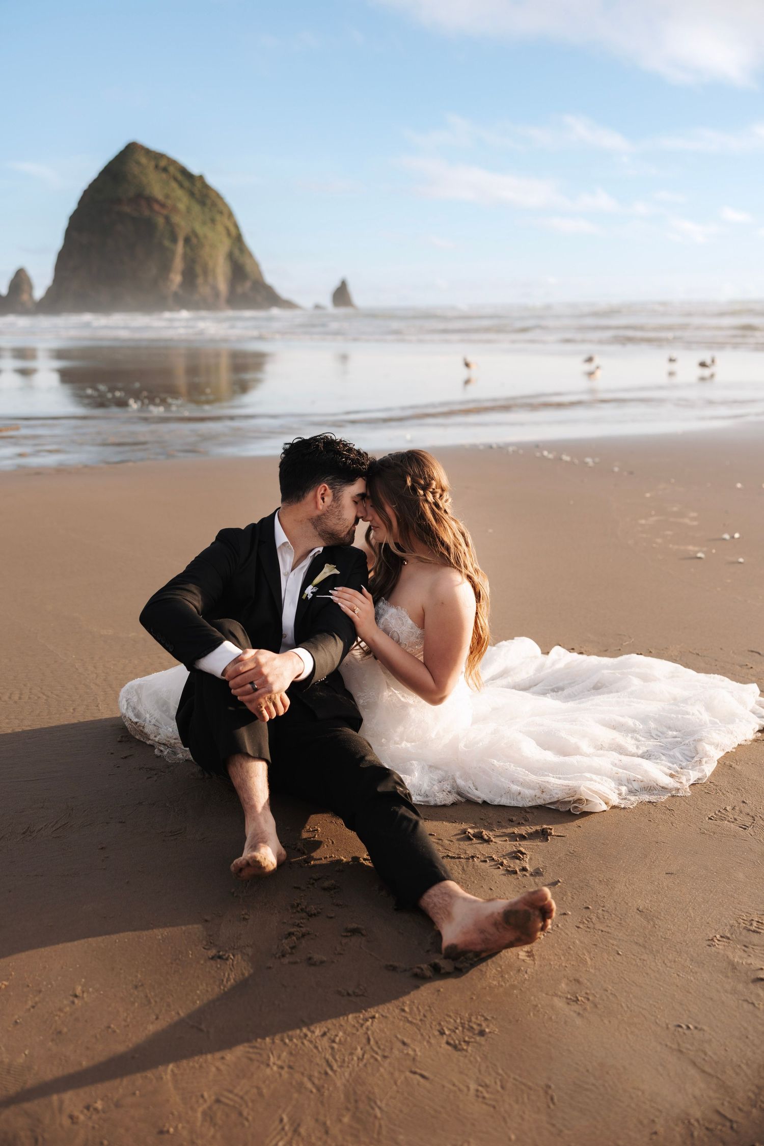 Barefoot bride and groom sitting together on beach sand with iconic Haystack Rock in background.