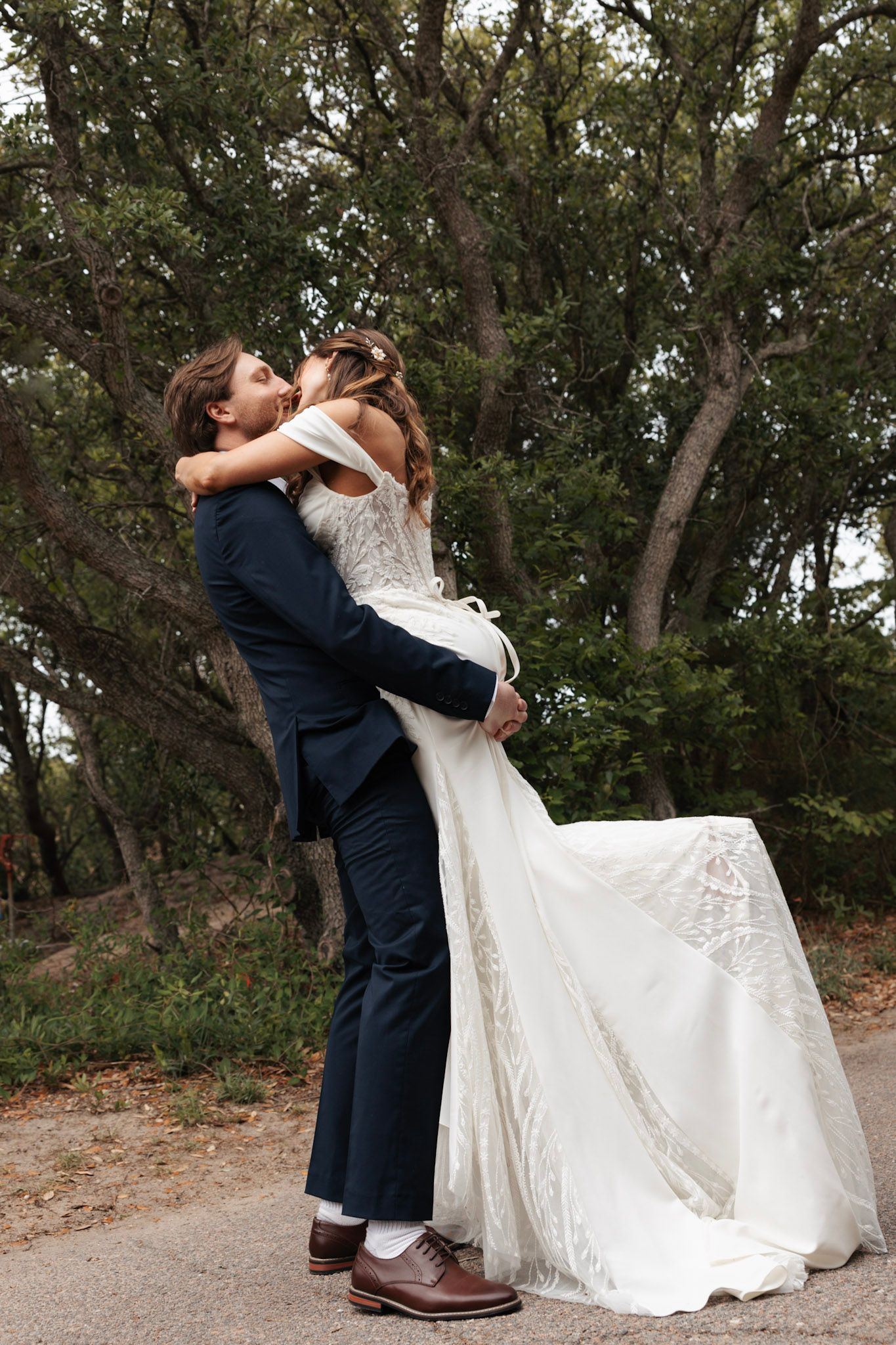 Groom lifting and kissing bride in lace wedding gown among lush green trees in outdoor setting.