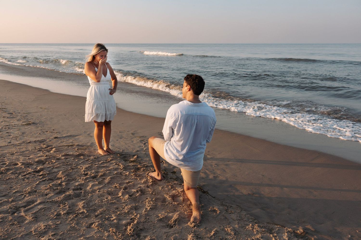 Man proposing on one knee to woman in white dress on sandy beach at sunset.